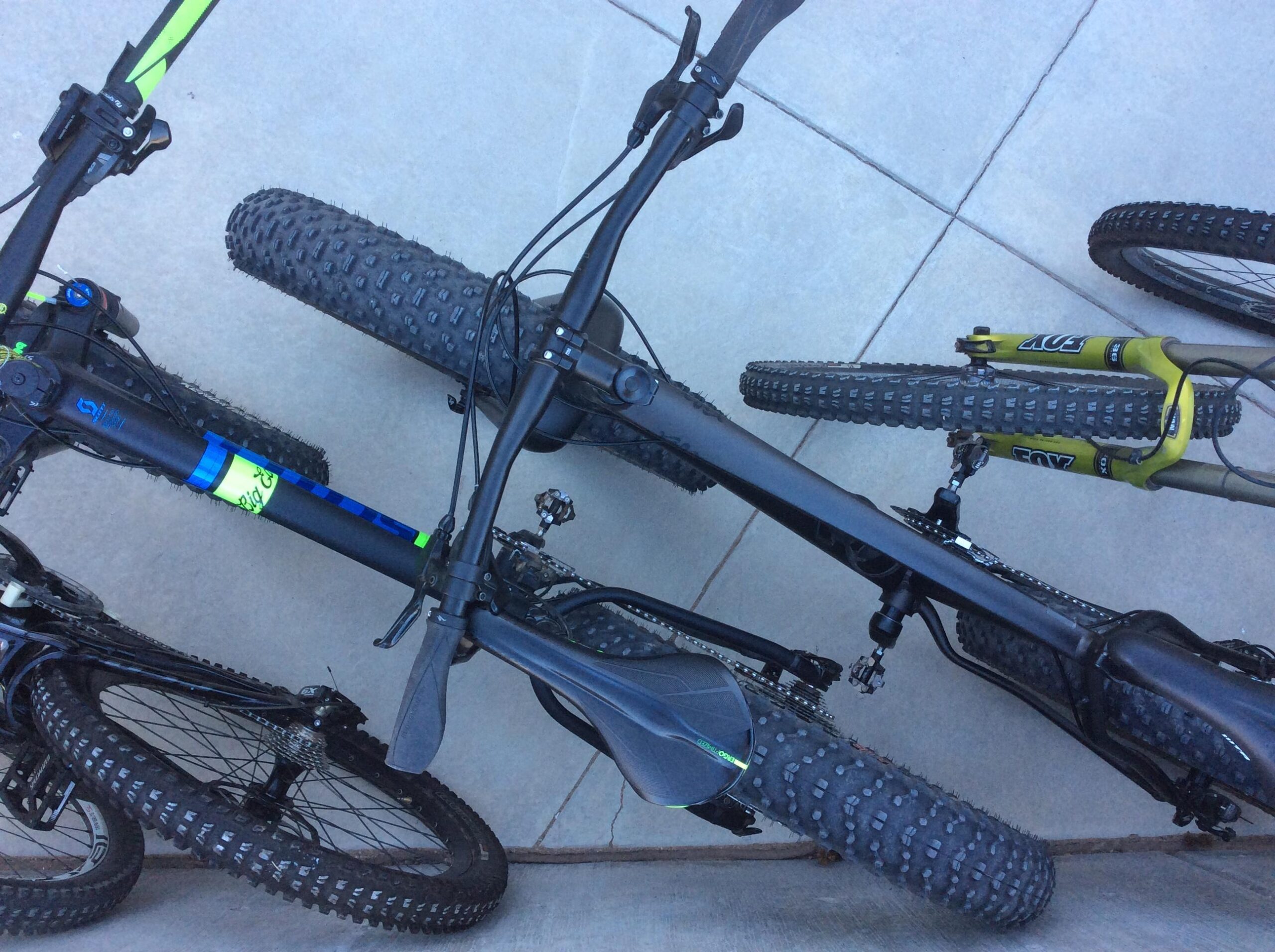 Two mountain bikes are positioned closely together, showing their frames, handlebars, and knobby tires. The bikes have a mix of black and bright accents, with one featuring a green and blue design. The background is a light-colored concrete surface.