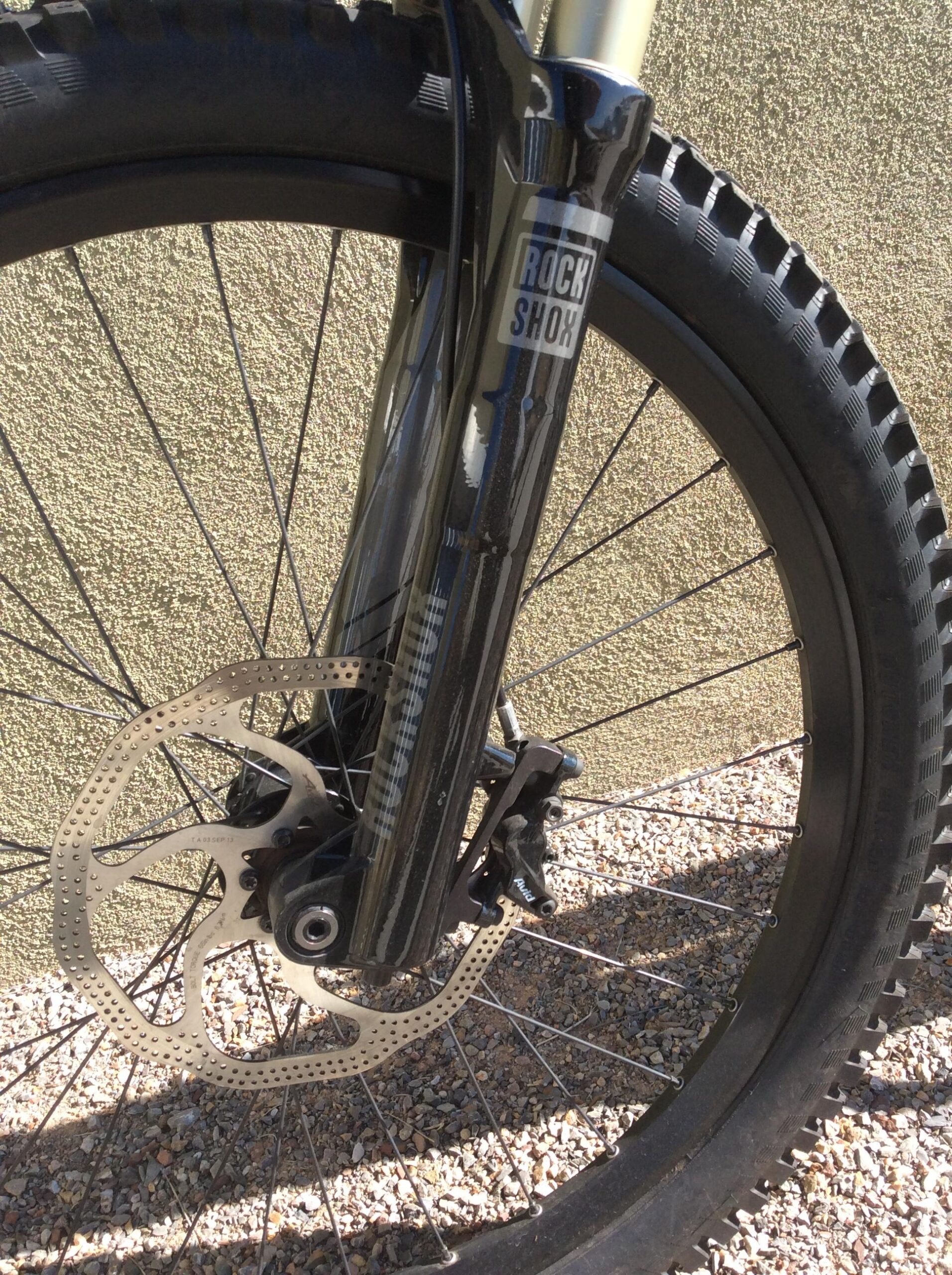Specialized S-Works Demo 8: Close-up image of a mountain bike front fork, featuring a RockShox brand suspension fork and a disc brake system. The bike's wheel, tire, and part of the gravel surface are visible in the background.