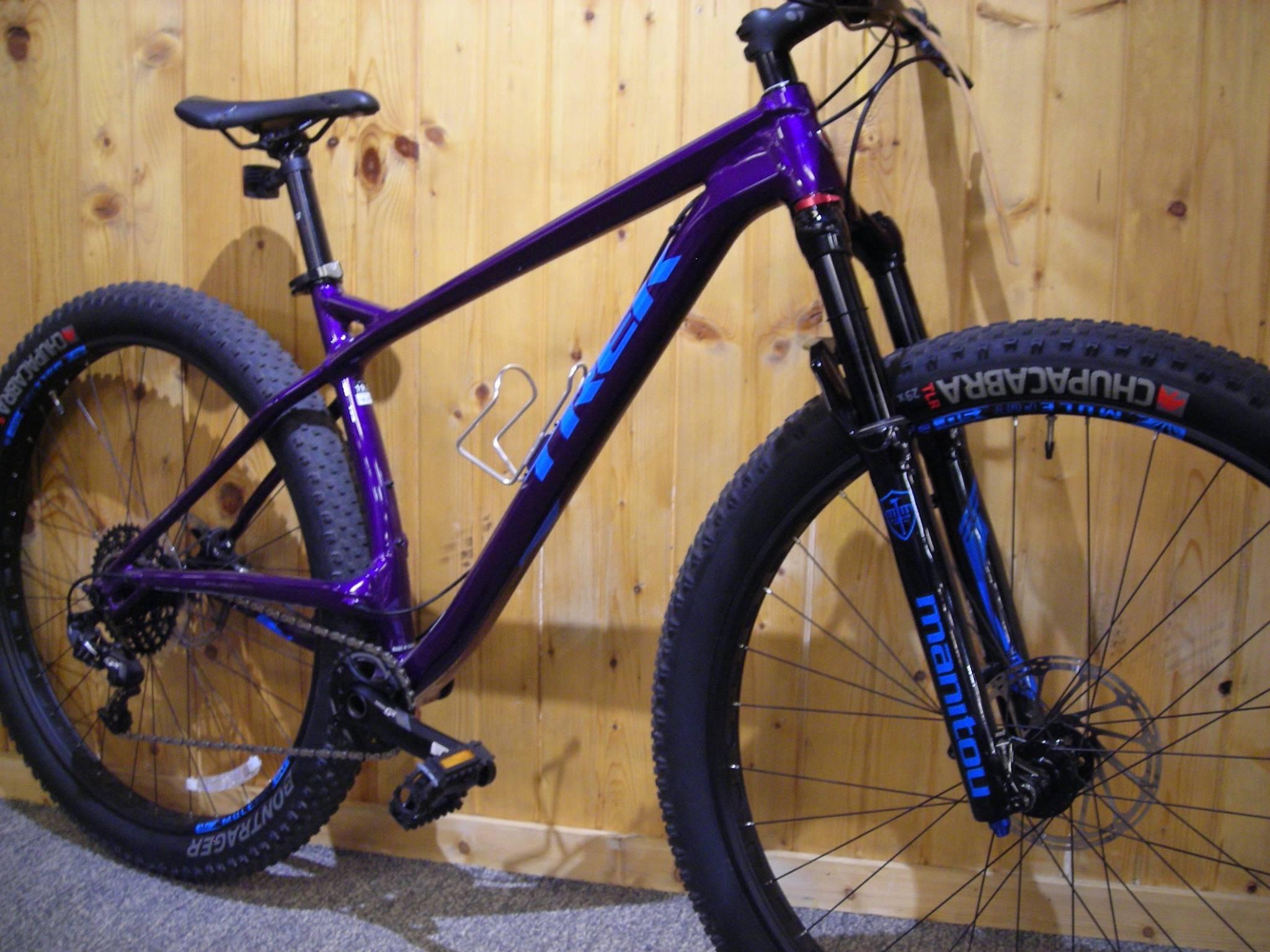 Trek Stache: A vibrant purple mountain bike leaning against a wooden wall, showcasing its sleek design, wide tires, and high-performance components. The bike features a black front fork with blue lettering, adjustable seat post, and detailed drivetrain, emphasizing its suitability for off-road adventures.