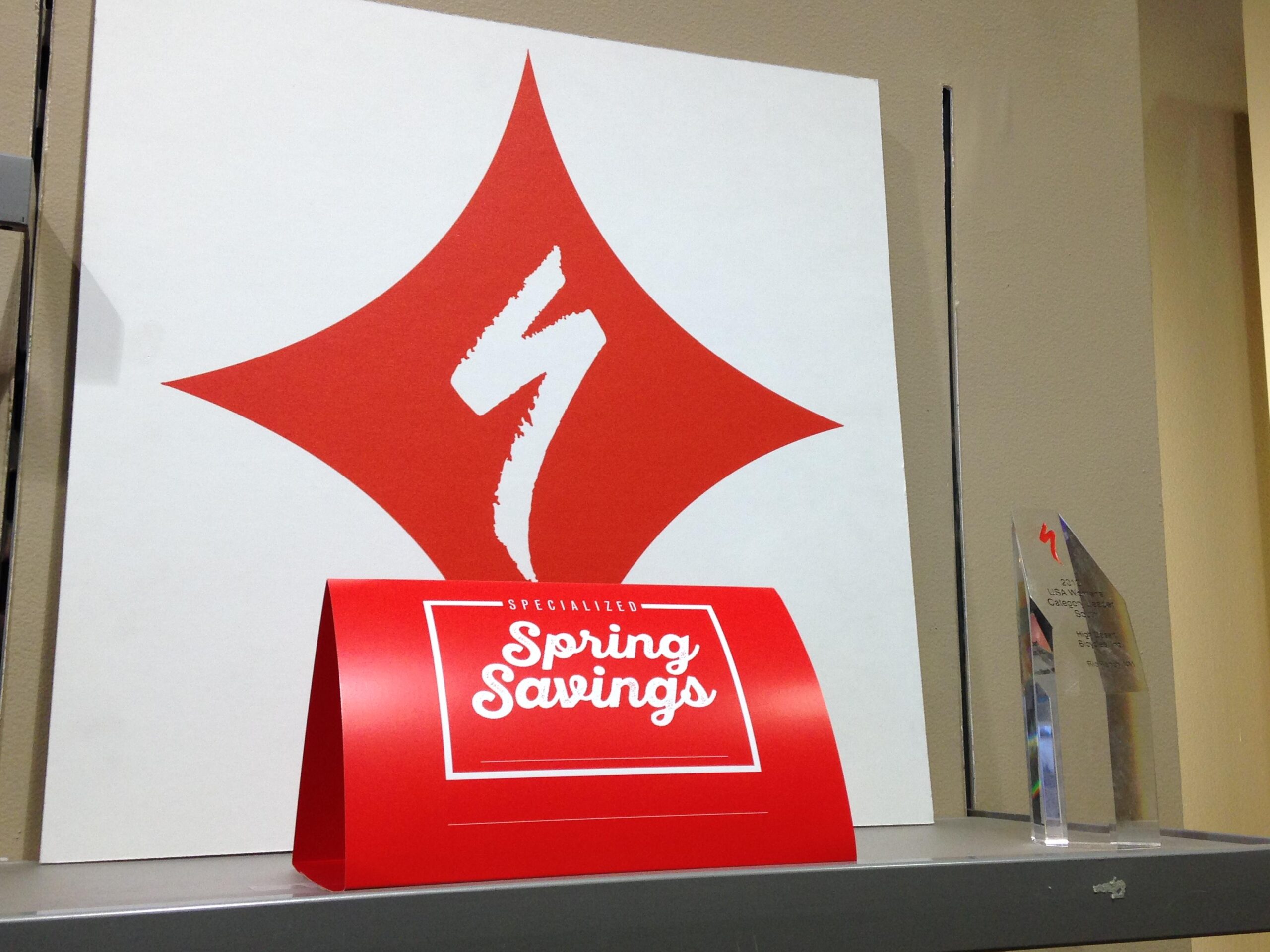 A display featuring a large white poster with a red diamond shape and a stylized "S" logo, next to a bright red promotional stand labeled "Spring Savings," and an award trophy on a shelf.