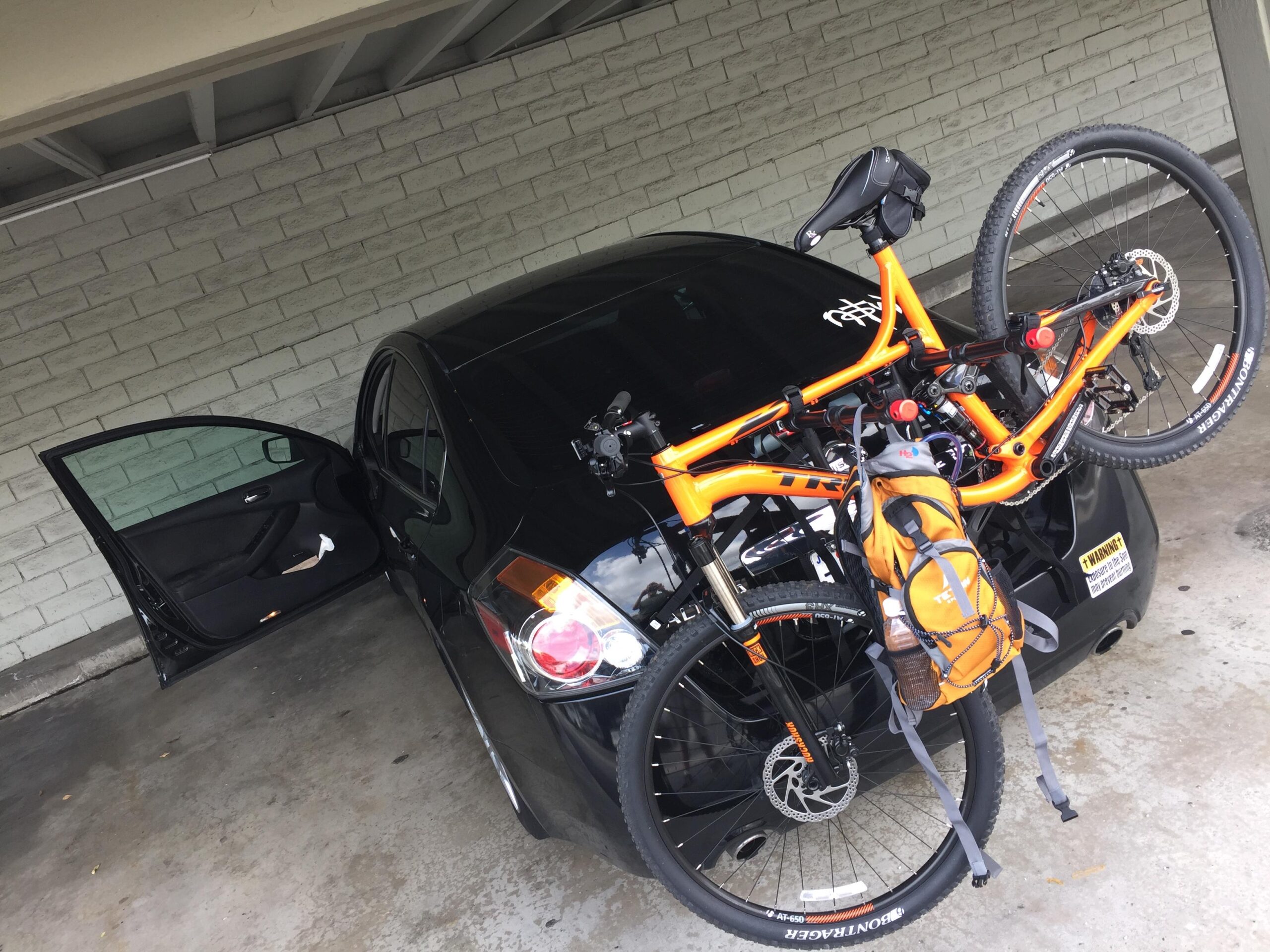 Trek Fuel EX 5 29: A black car parked under a covered area with the driver's side door open. An orange mountain bike is mounted on the back of the car, with a small backpack attached to it. The background features a light-colored brick wall.
