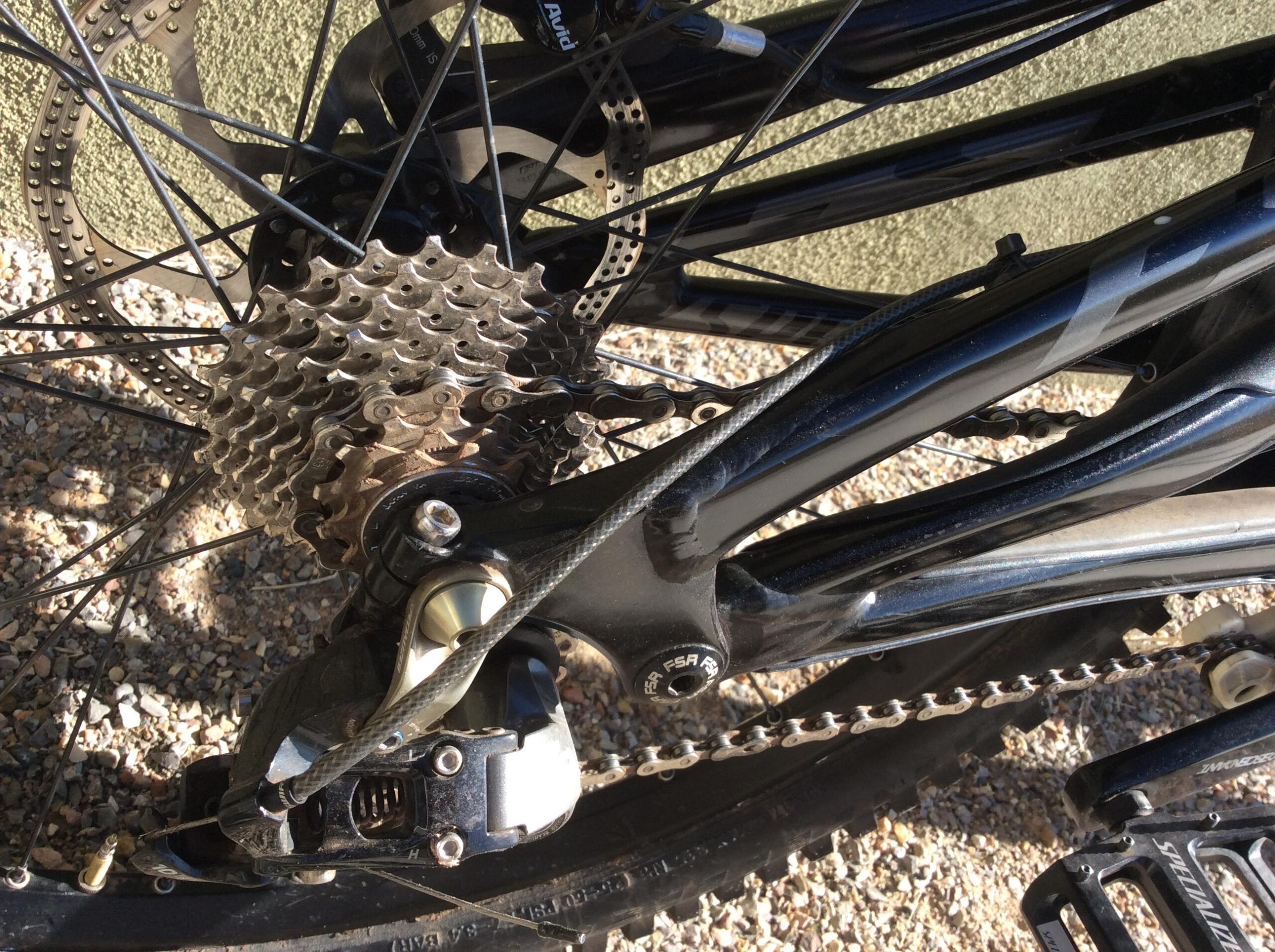 Specialized S-Works Demo 8: Close-up of a bicycle's rear derailleur, chain, and cassette with a black frame set against a gravel background.