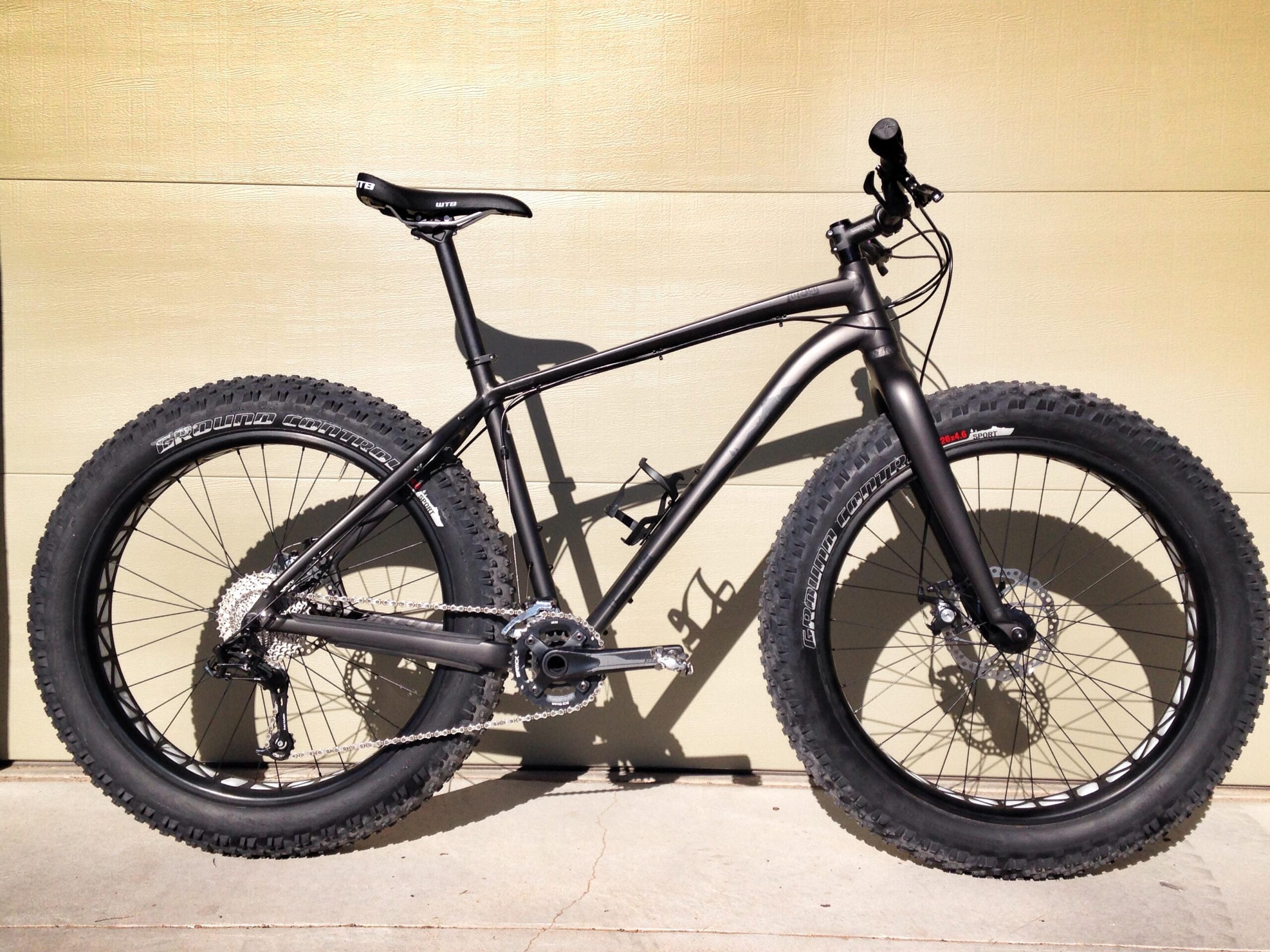 A black fat bike with oversized tires is positioned against a light-colored wall. The bike features a sturdy frame, disc brakes, and a single gear setup, highlighting its design for rough terrain and off-road biking. The tires have a prominent tread pattern for enhanced grip on various surfaces.