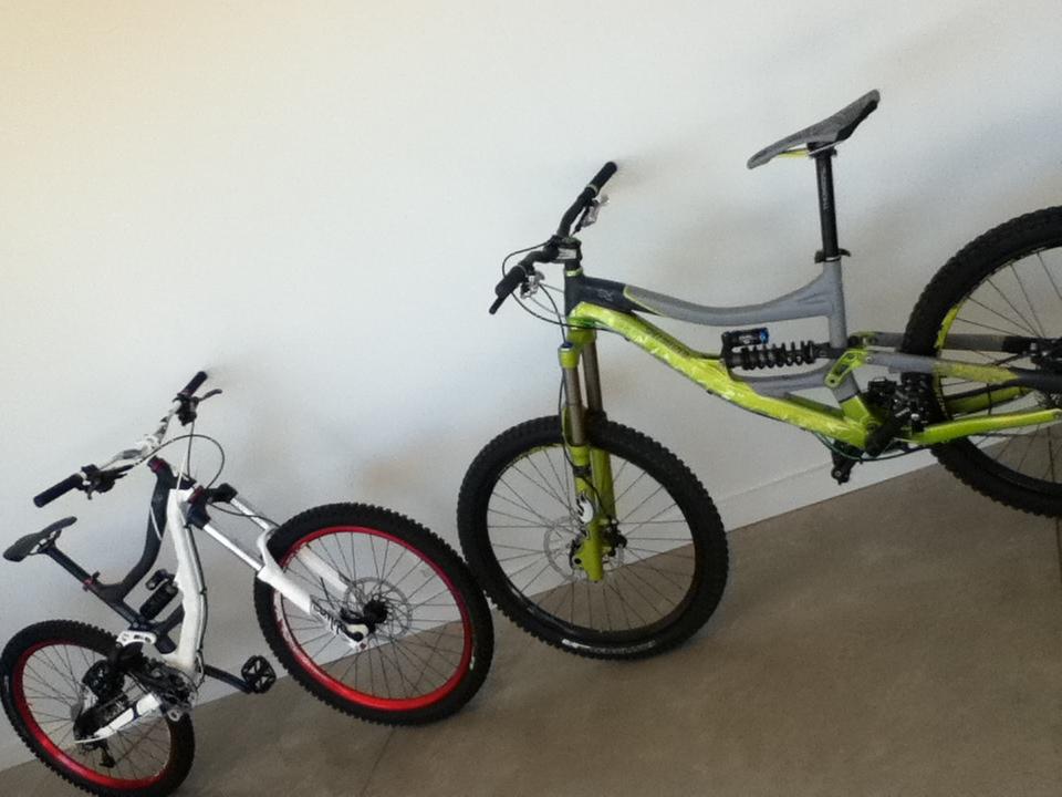 Two mountain bikes are leaning against a light-colored wall. The bike on the left is predominantly white with red rims, while the bike on the right features a green and gray design, equipped with a shock absorber. Both bikes have thick tires suitable for off-road cycling, and the setting has a minimalist vibe with a concrete floor.