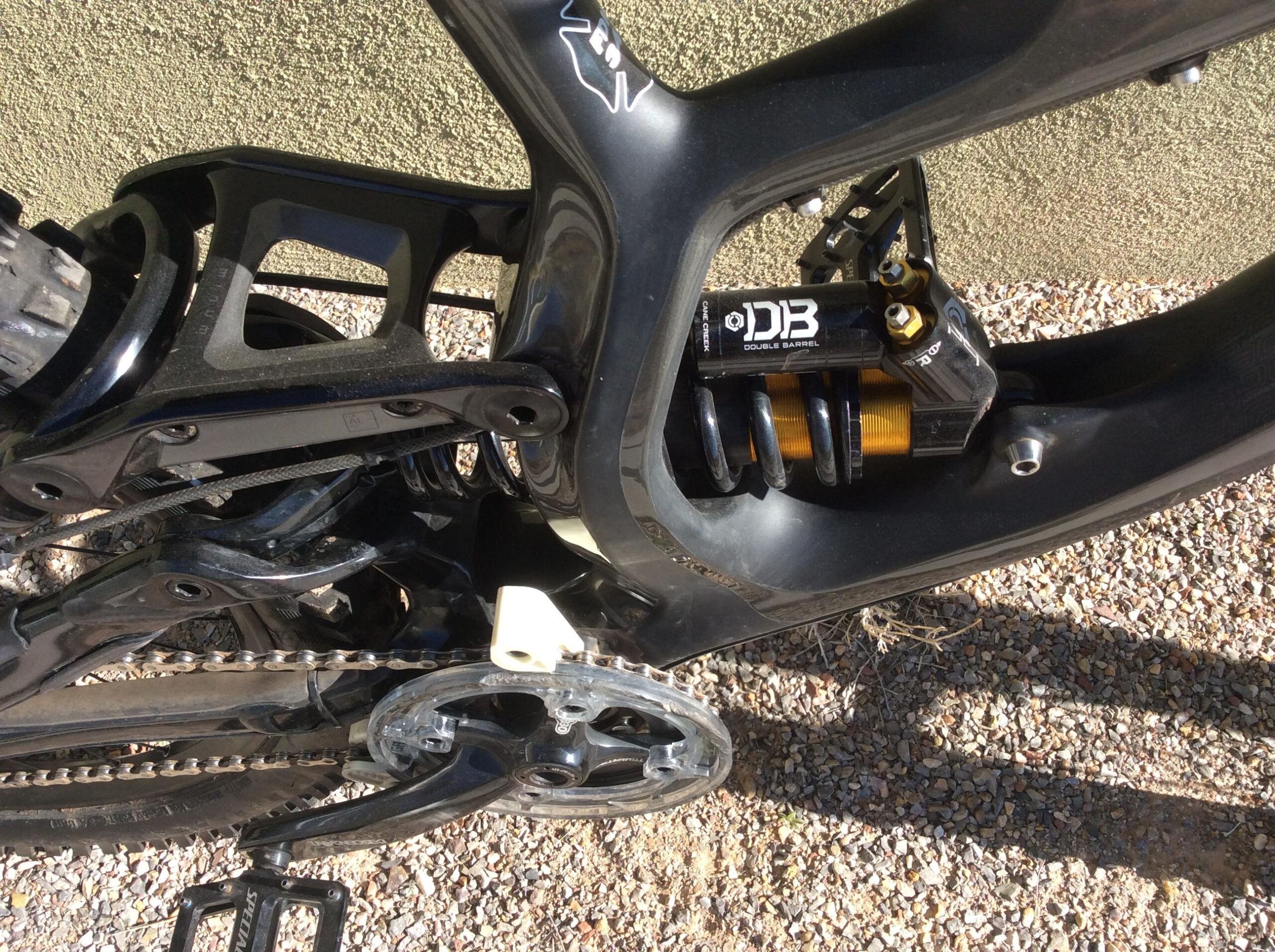 Specialized S-Works Demo 8: Close-up of a mountain bike's rear suspension system, showcasing the black frame, a visible shock absorber marked "DB," and the chain and crankset. The background features a gravel surface.