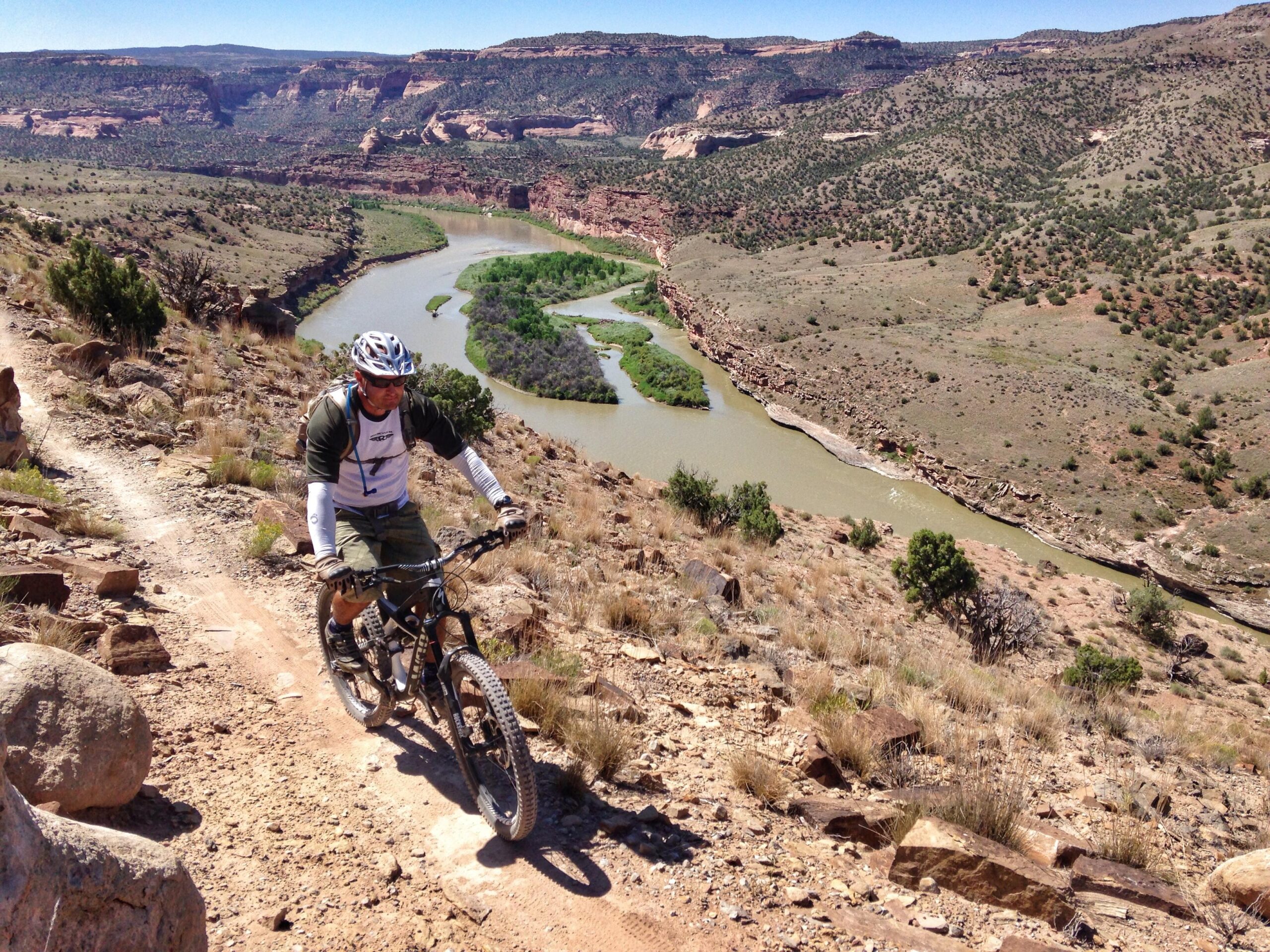 Specialized S-Works Enduro SL Carbon: A mountain biker riding along a dirt trail on a rocky hilltop, overlooking a winding river and green vegetation below, with distant hills and blue sky in the background.