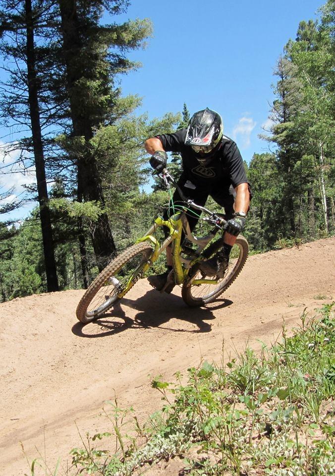A mountain biker in a black shirt and helmet leans into a turn while riding a bright green bike on a dirt trail surrounded by tall trees and clear blue skies. Angel Fire Bike Park mountain bike trail.