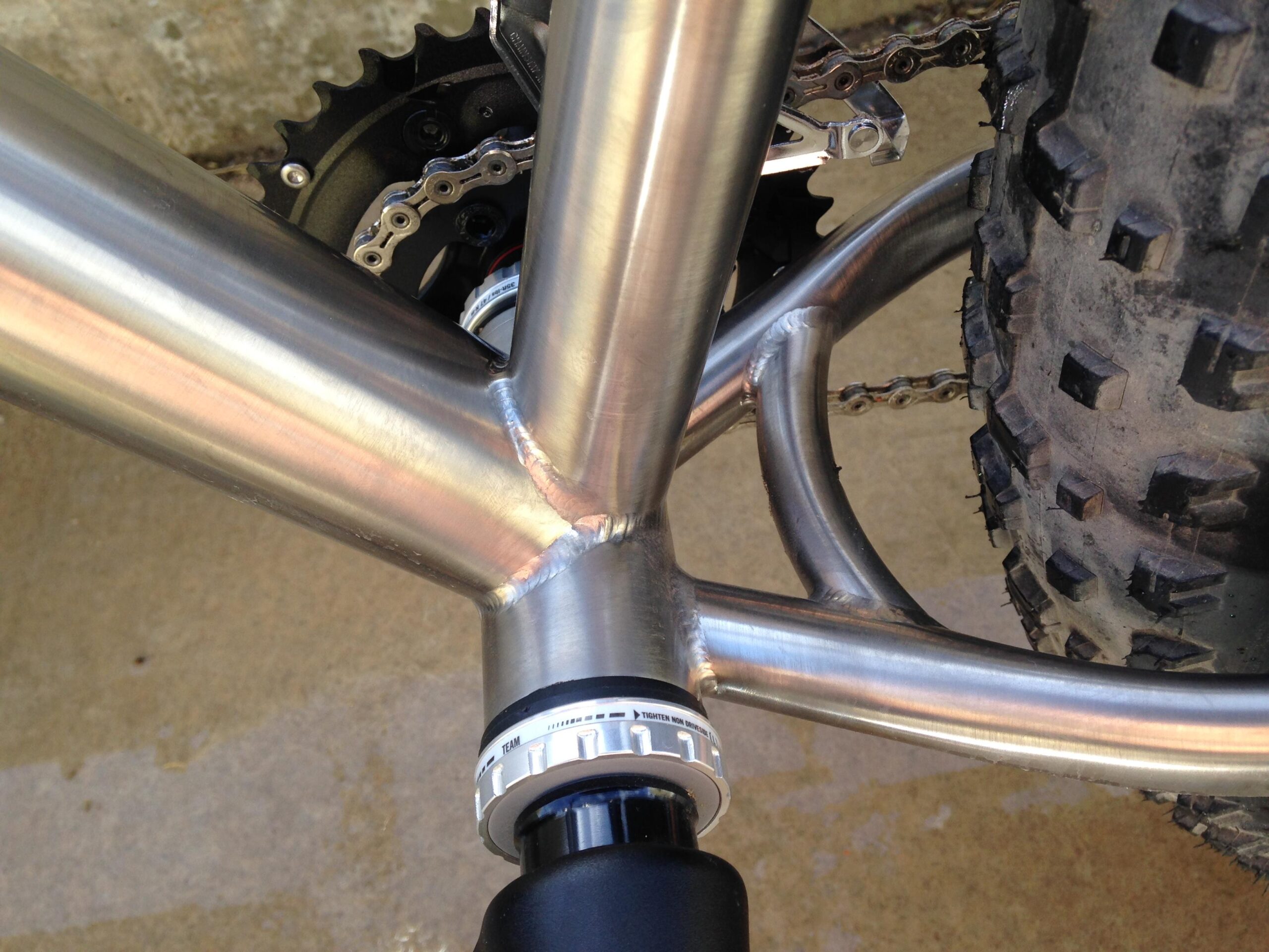 Motobecane Motobecane Bullet Titanium Fat bike: Close-up view of a bicycle's bottom bracket area, showcasing the welded stainless steel frame, gear assembly, and part of the tire. The image emphasizes the craftsmanship of the frame construction and the components involved.