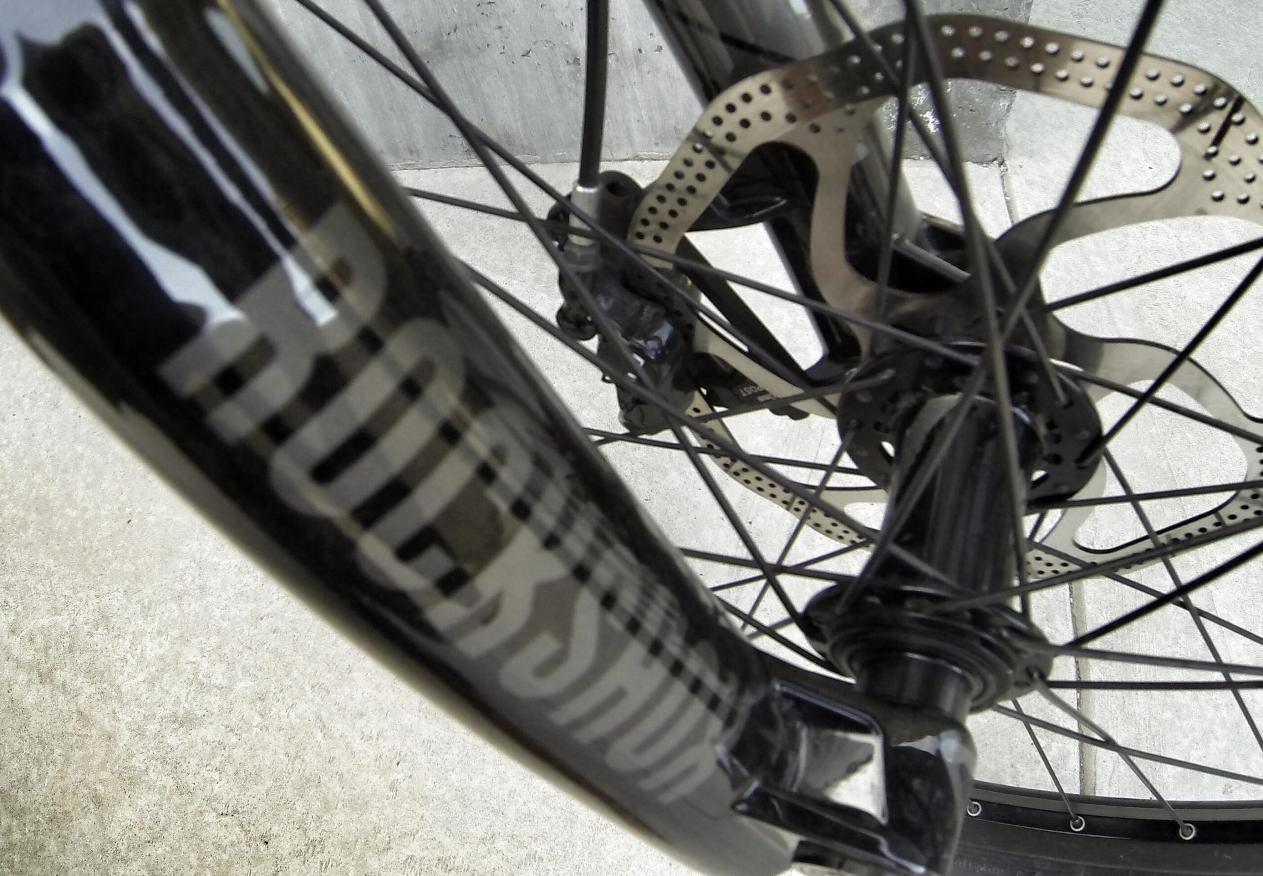 Specialized S-Works Demo 8: Close-up view of a bike's front wheel, featuring the RockShox logo on the fork, with a detailed look at the disc brake and spokes. The background shows a concrete surface.
