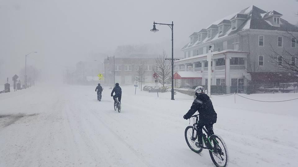 Three cyclists ride on a snow-covered street during a heavy snowstorm, with visibility heavily reduced due to falling snow. A building can be seen in the background along with street lamps and traffic signs. The scene captures the challenges of biking in winter weather. Lewis Morris mountain bike trail.