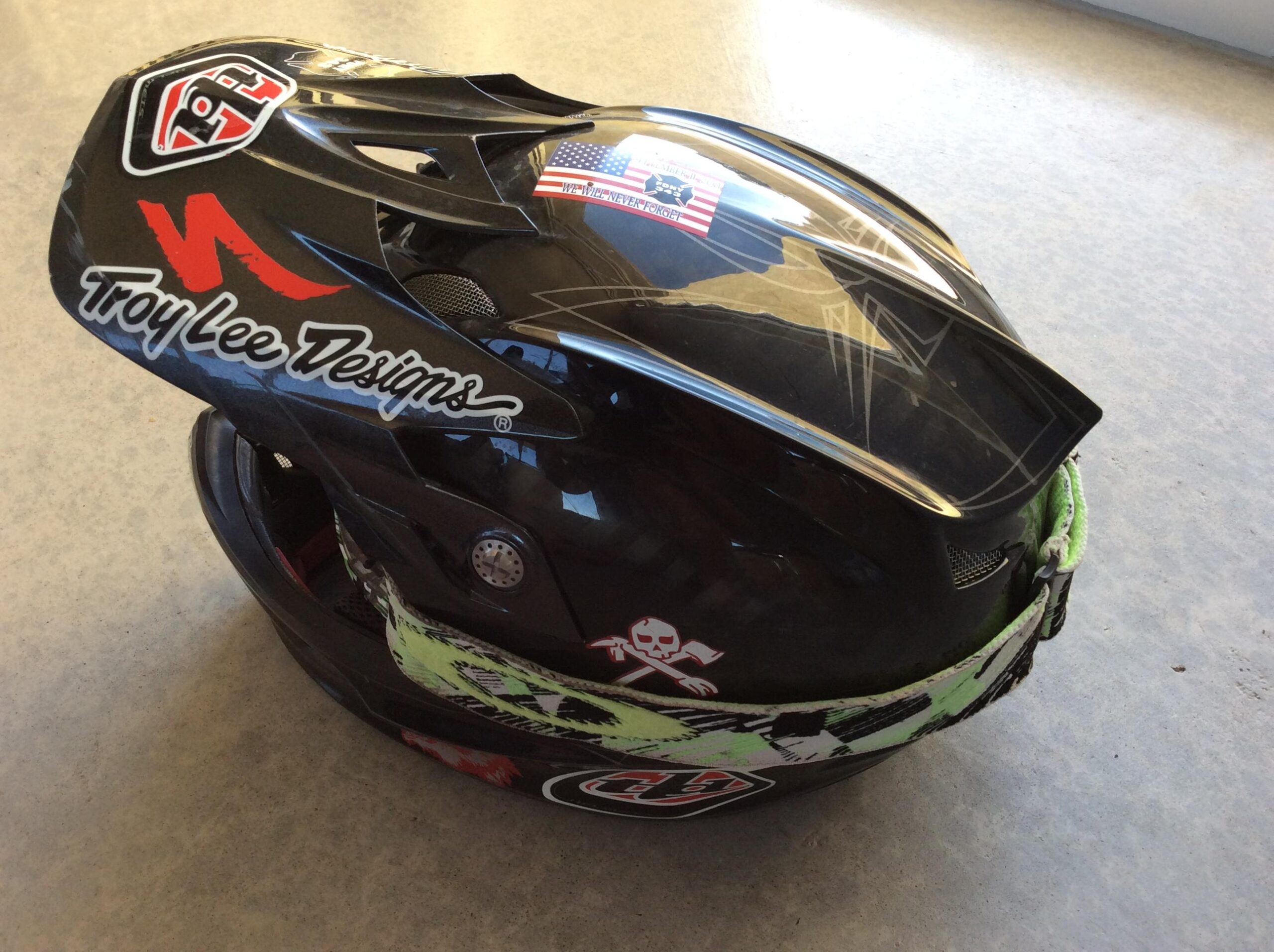 Alt text: A black helmet featuring the logo "Troy Lee Designs," with decorative patterns and stickers, including an American flag and a skull design, placed on a flat surface.