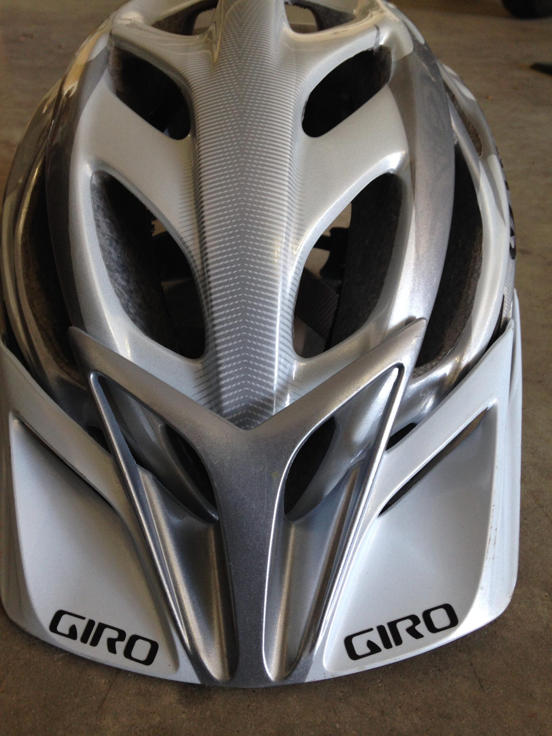 Close-up view of a silver Giro cycling helmet, showcasing its sleek design and ventilation openings. The helmet features a distinctive aerodynamic shape with a textured top and the Giro logo prominently displayed on the sides. The background is a neutral surface, highlighting the helmet's details.