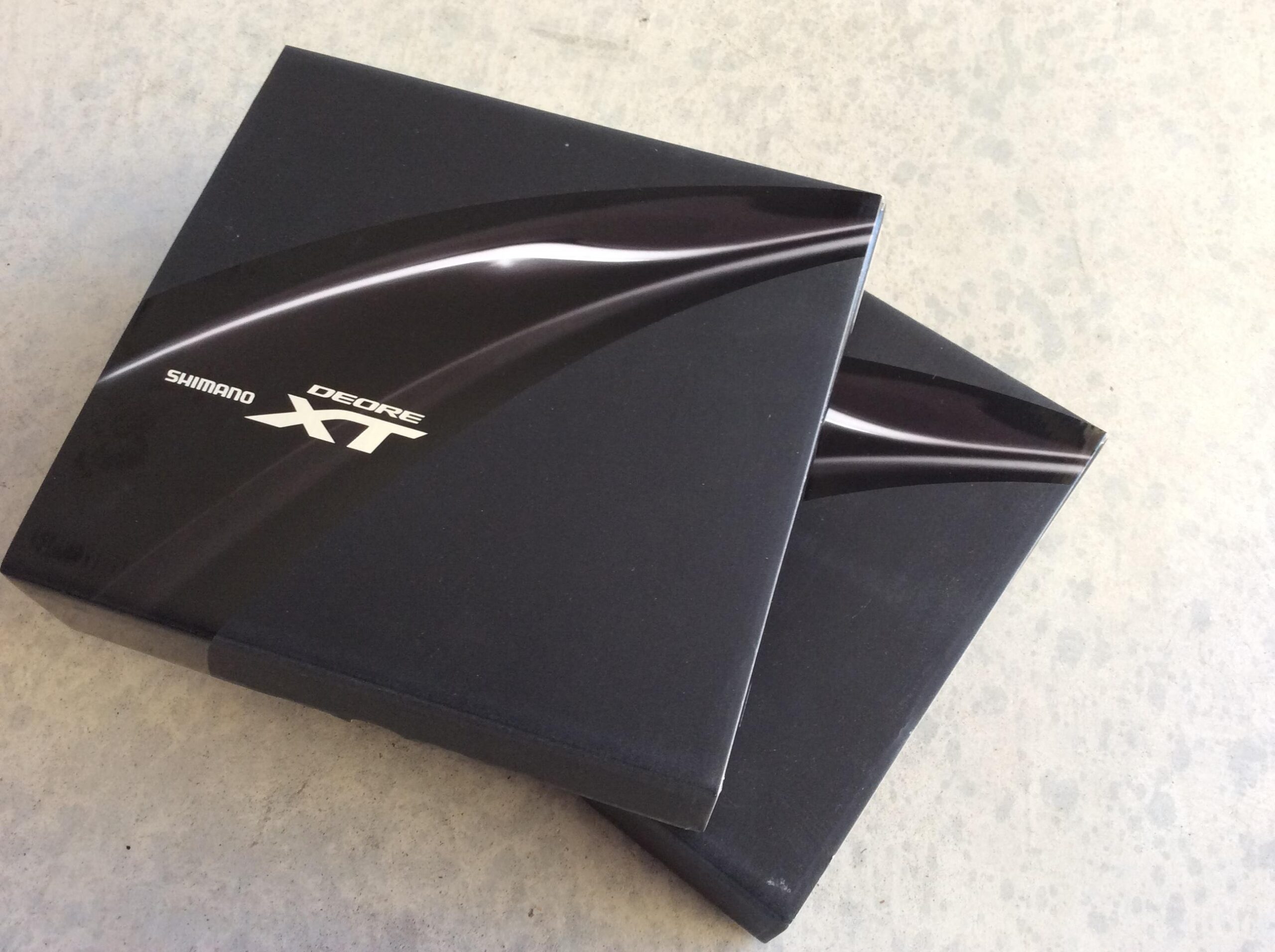Two Shimano Deore XT boxes stacked on top of each other, featuring a sleek black design with a subtle glossy line pattern and the Shimano logo prominently displayed. The boxes rest on a gray concrete surface.