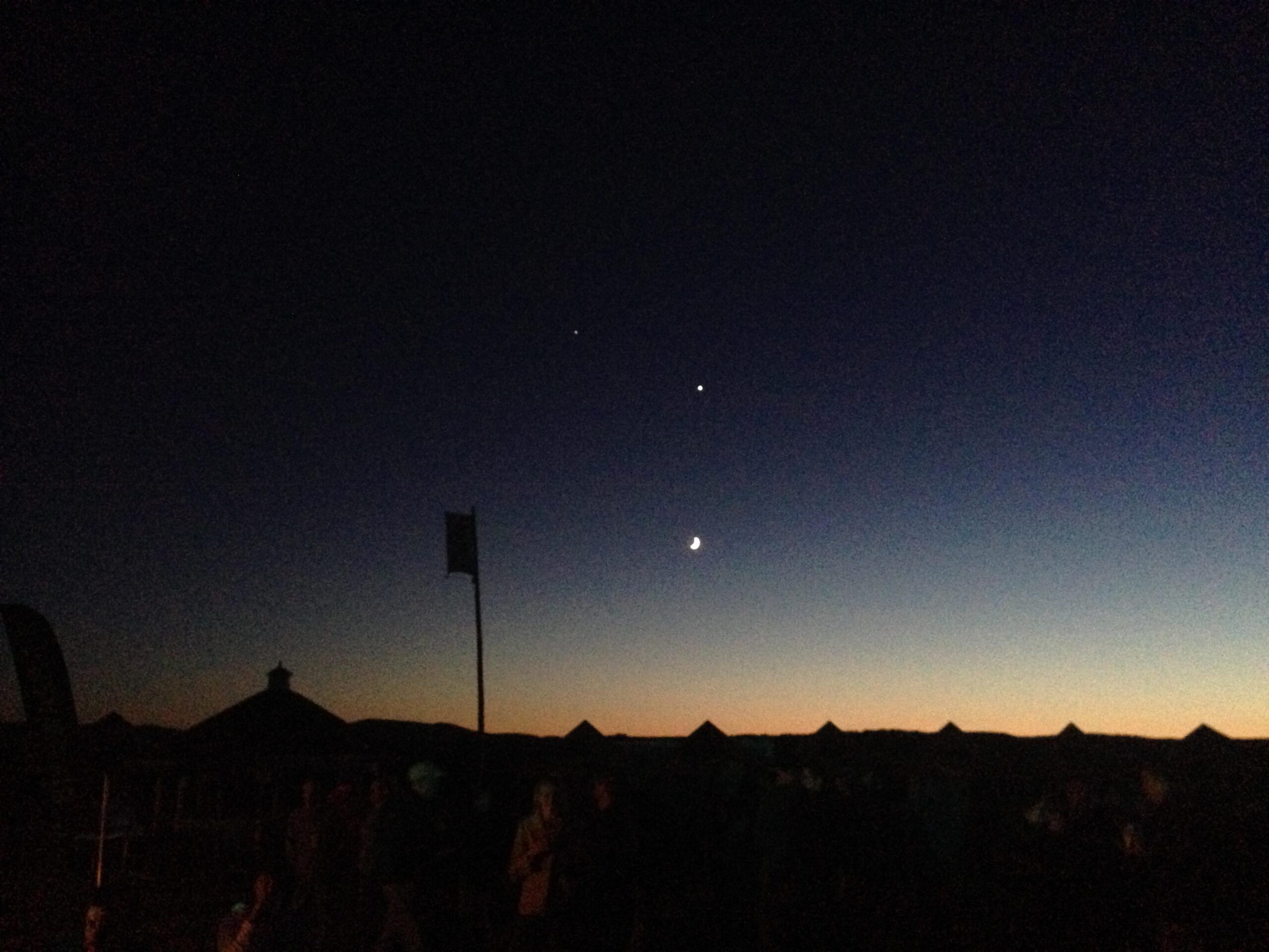 A twilight sky transitioning from deep blue to lighter hues at the horizon, featuring a crescent moon and two bright celestial bodies. Silhouetted figures can be seen gathering below, along with distant dark shapes of structures. Kingdom Trails mountain bike trail.