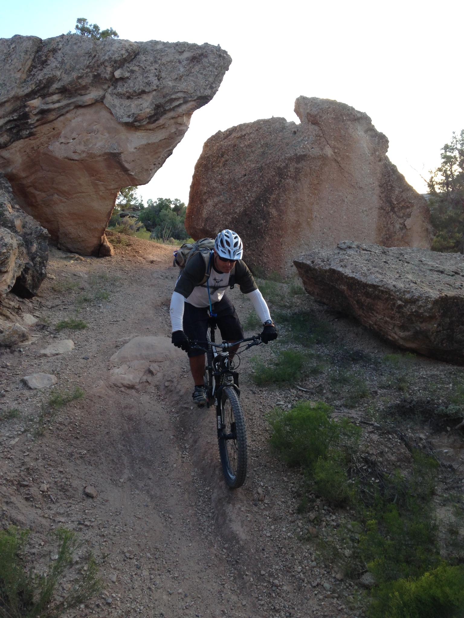 Specialized Enduro S-Works 650b: A mountain biker navigating a rocky terrain, flanked by large boulders and surrounded by sparse vegetation. The sun is setting in the background, casting a warm glow on the landscape. The rider is wearing a helmet and protective gear.
