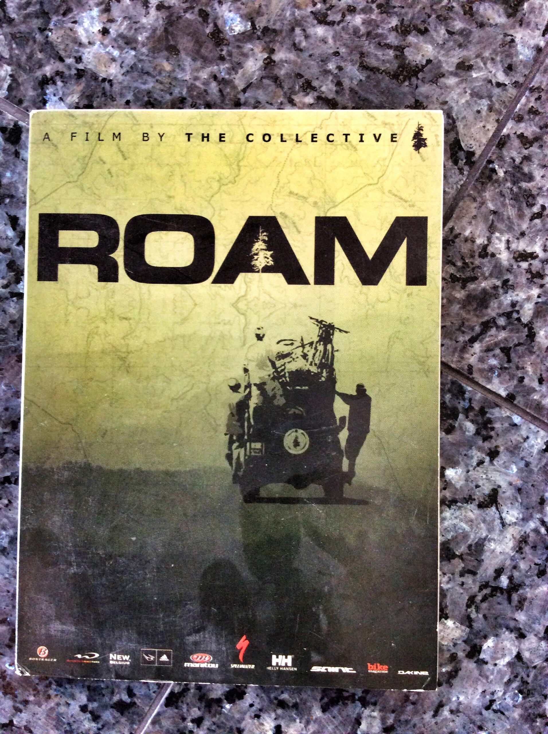 A DVD cover for the film "ROAM," featuring a green background with a silhouette of a vehicle carrying gear, accompanied by two figures. The title "ROAM" is prominently displayed in bold black letters, with smaller text indicating it is a film by "THE COLLECTIVE." The background includes a subtle map design.