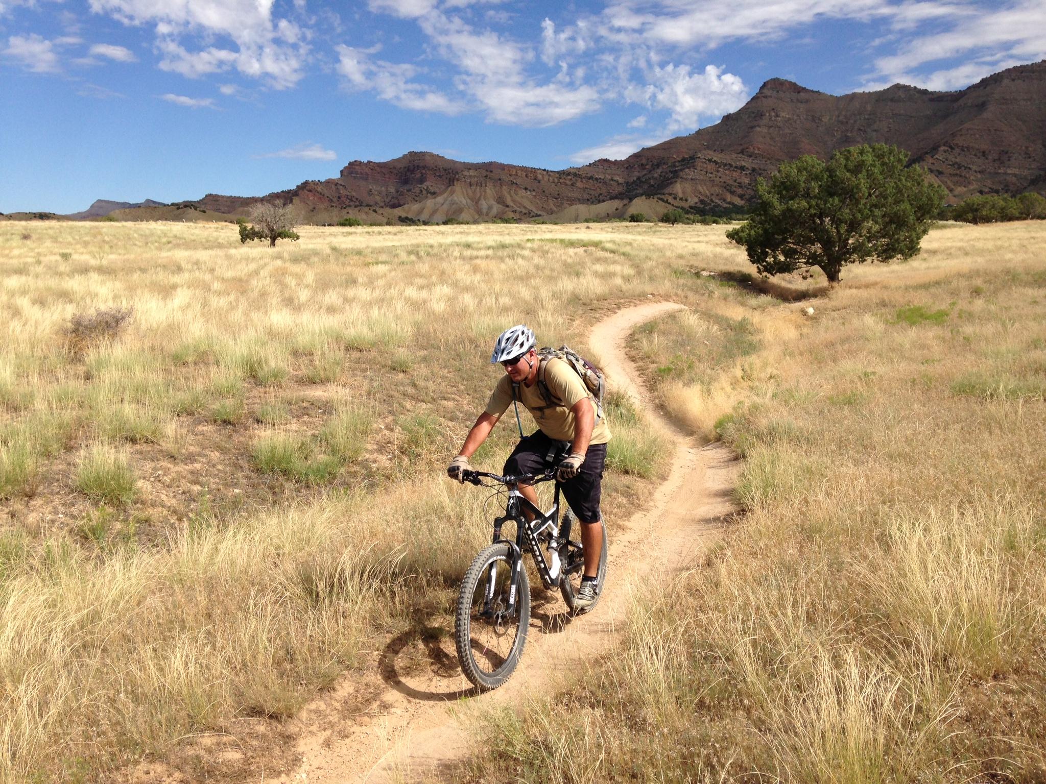 A mountain biker riding on a dirt path through a grassy field, with rugged hills and blue skies in the background. 18 Road Trails / North Fruita Desert mountain bike trail.