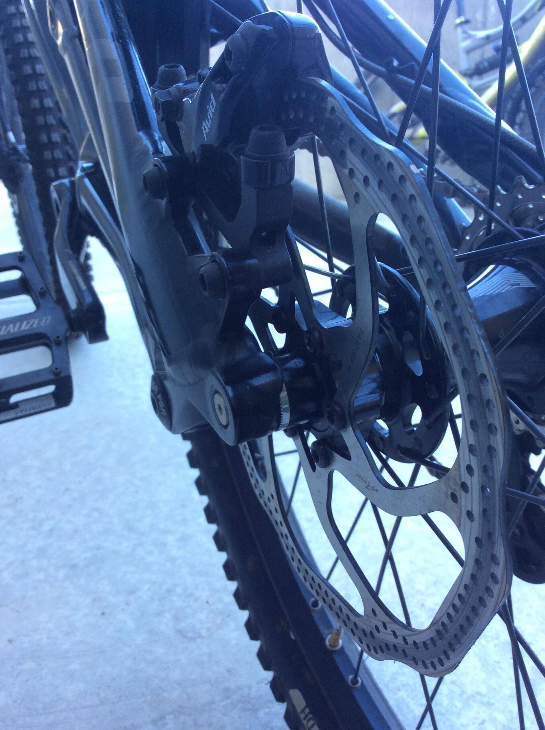 Specialized S-Works Demo 8 Carbon: Close-up view of a mountain bike's rear brake assembly, showcasing the brake caliper, rotor, and part of the bike frame and pedal. The components are primarily black with a silver rotor featuring holes for weight reduction.