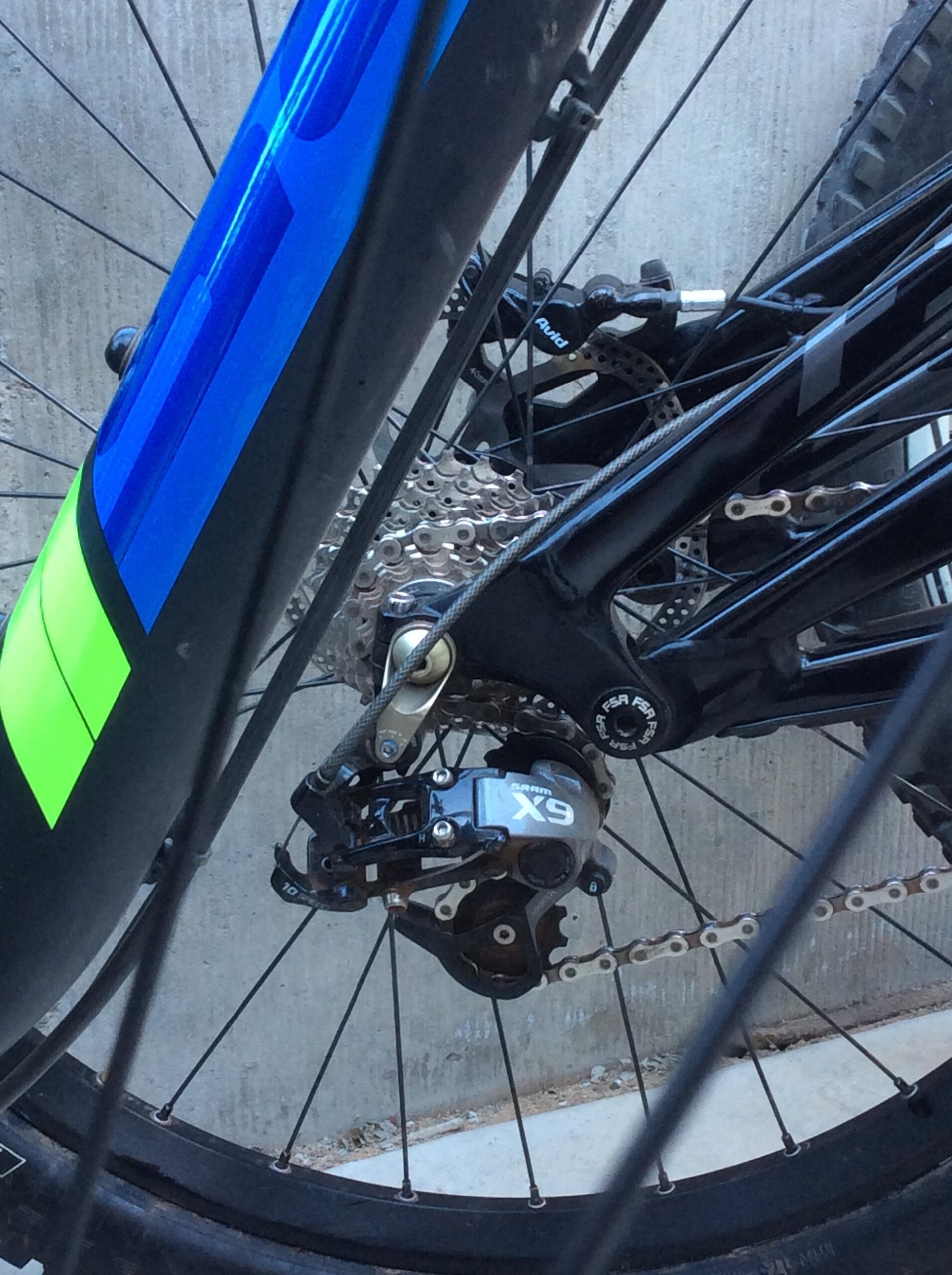 Close-up of a bicycle rear derailleur and cassette, showcasing the SRAM X9 component. The bike frame features a blue and green design, with visible spokes and chain links. The background is a concrete surface.
