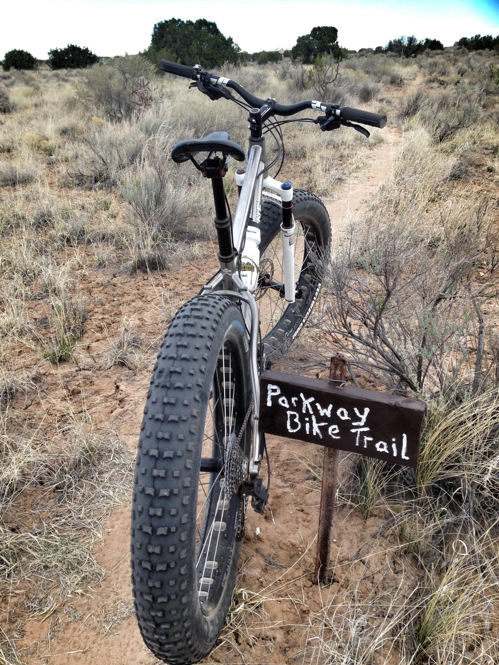 A mountain bike positioned on a dirt path in a grassy landscape, with a wooden sign reading "Parkway Bike Trail" nearby. The bike has thick, textured tires and a suspension system, suggesting it's ready for off-road riding. The background features shrubs and sparse trees under a cloudy sky. Parkway Fatbike trail mountain bike trail.