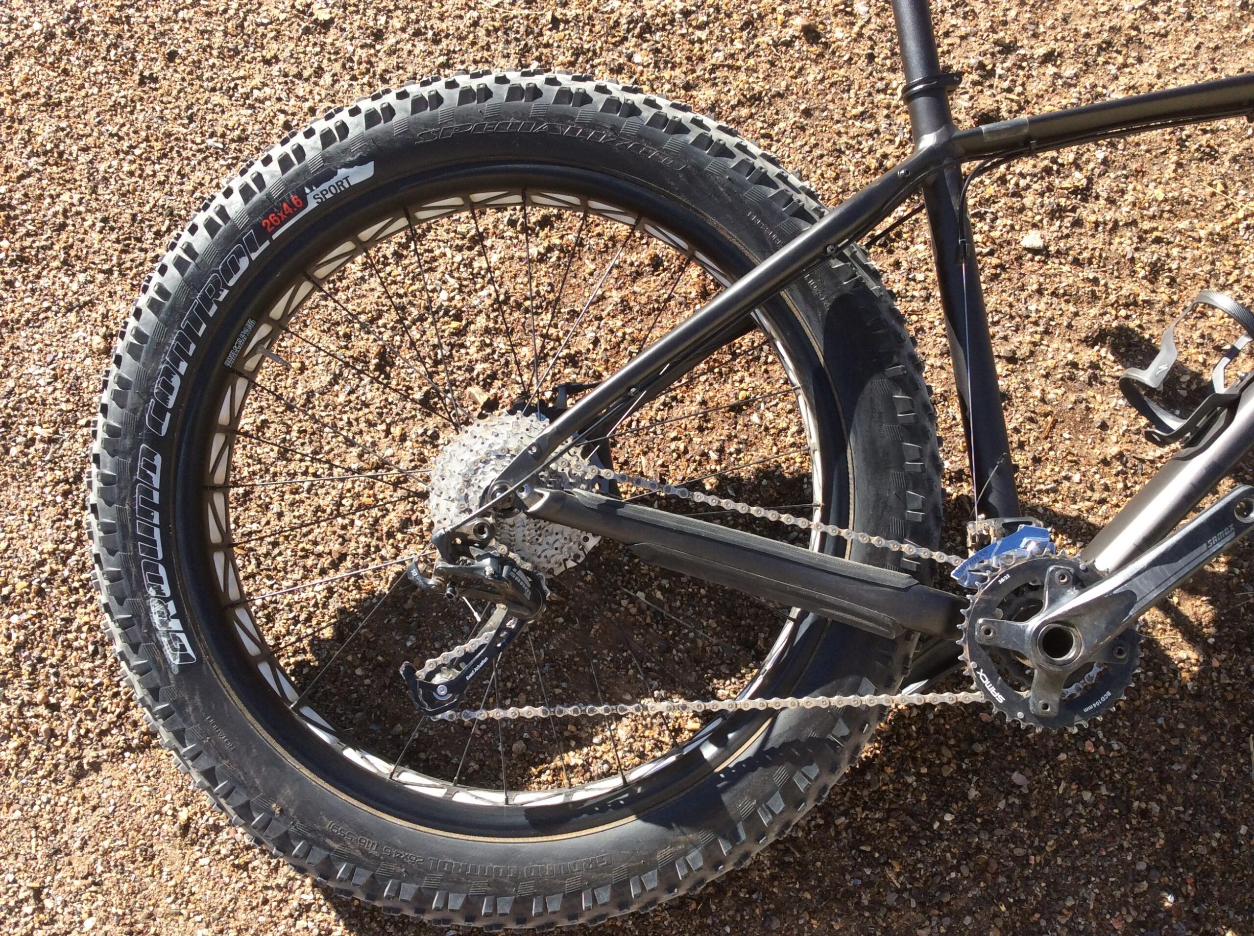 Specialized Fatboy: Close-up view of a mountain bike's rear wheel, featuring a knobby tire, gear cassette, and chain. The background consists of a gravel surface, highlighting the bike's rugged design suitable for off-road trails.