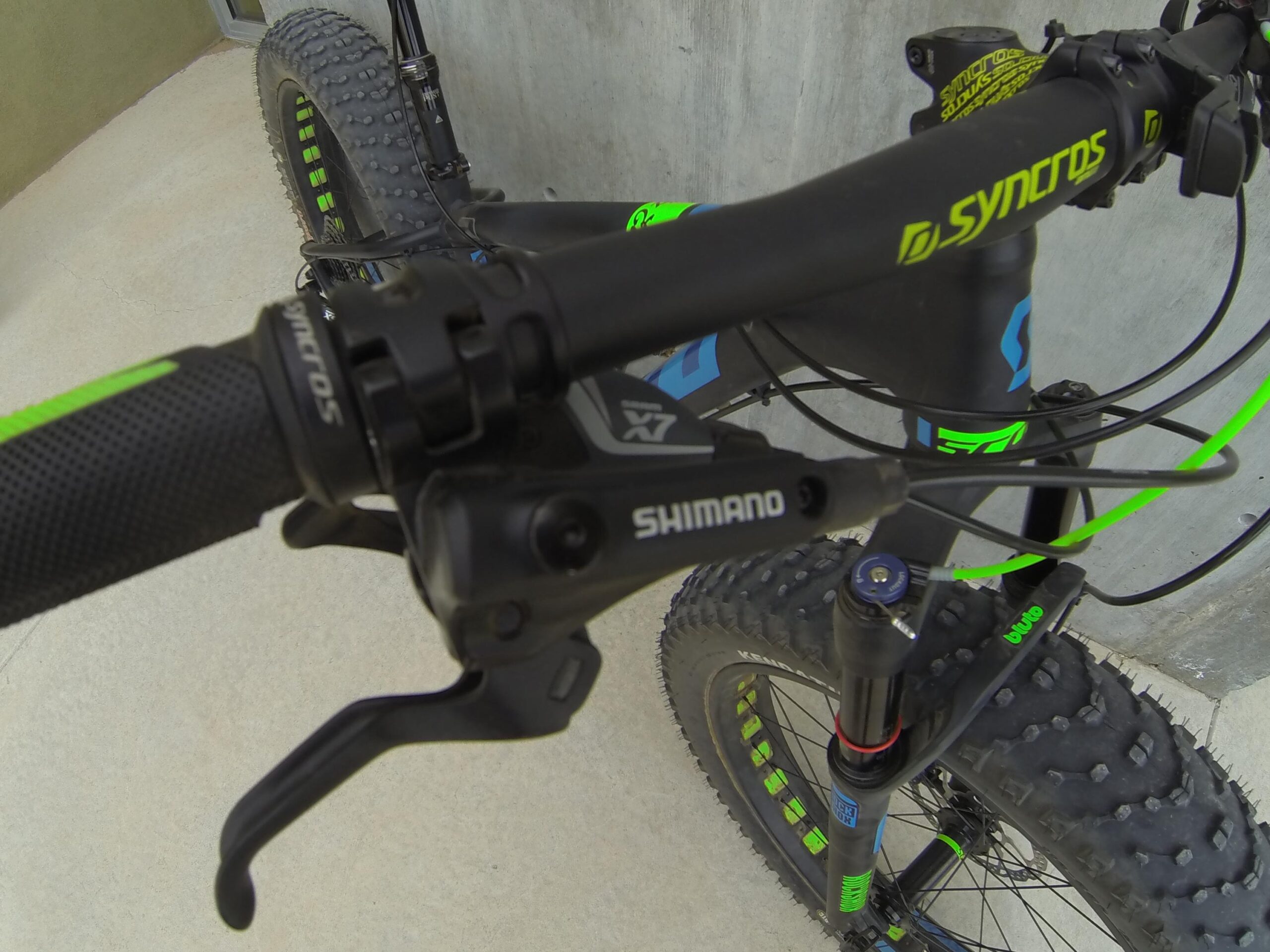 Scott Big Ed: Close-up view of a mountain bike handlebar featuring a Shimano brake lever, textured black grip, and bright green accents. The bike is equipped with chunky, knobby tires, emphasizing its suitability for rugged terrain. The background shows a concrete surface and a partially visible wall.