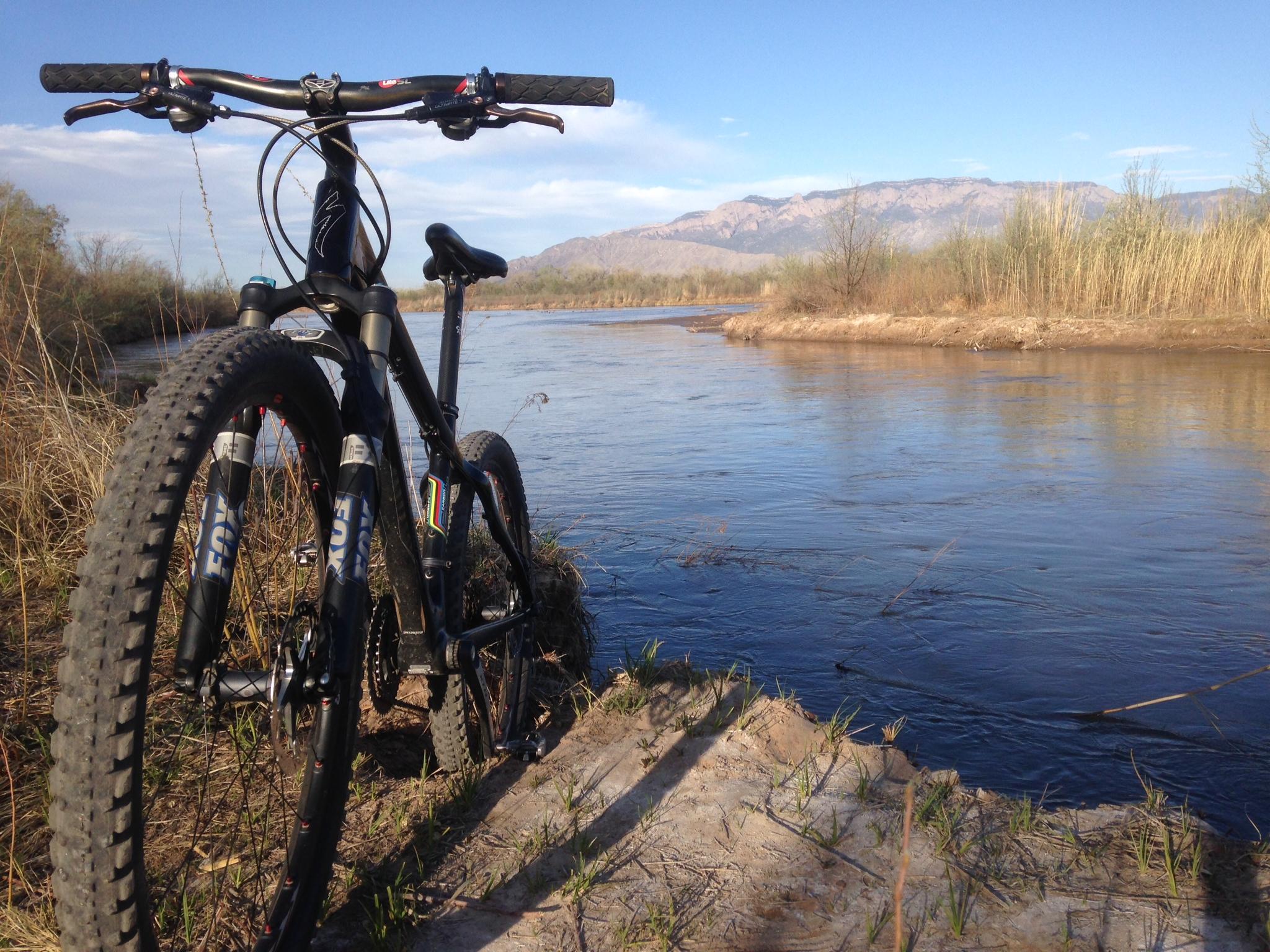 A mountain bike is positioned near a riverbank, with the water flowing gently in the background. The scene includes grassy areas and trees, under a clear blue sky with distant mountains visible on the horizon. Albuquerque Bosque mountain bike trail.