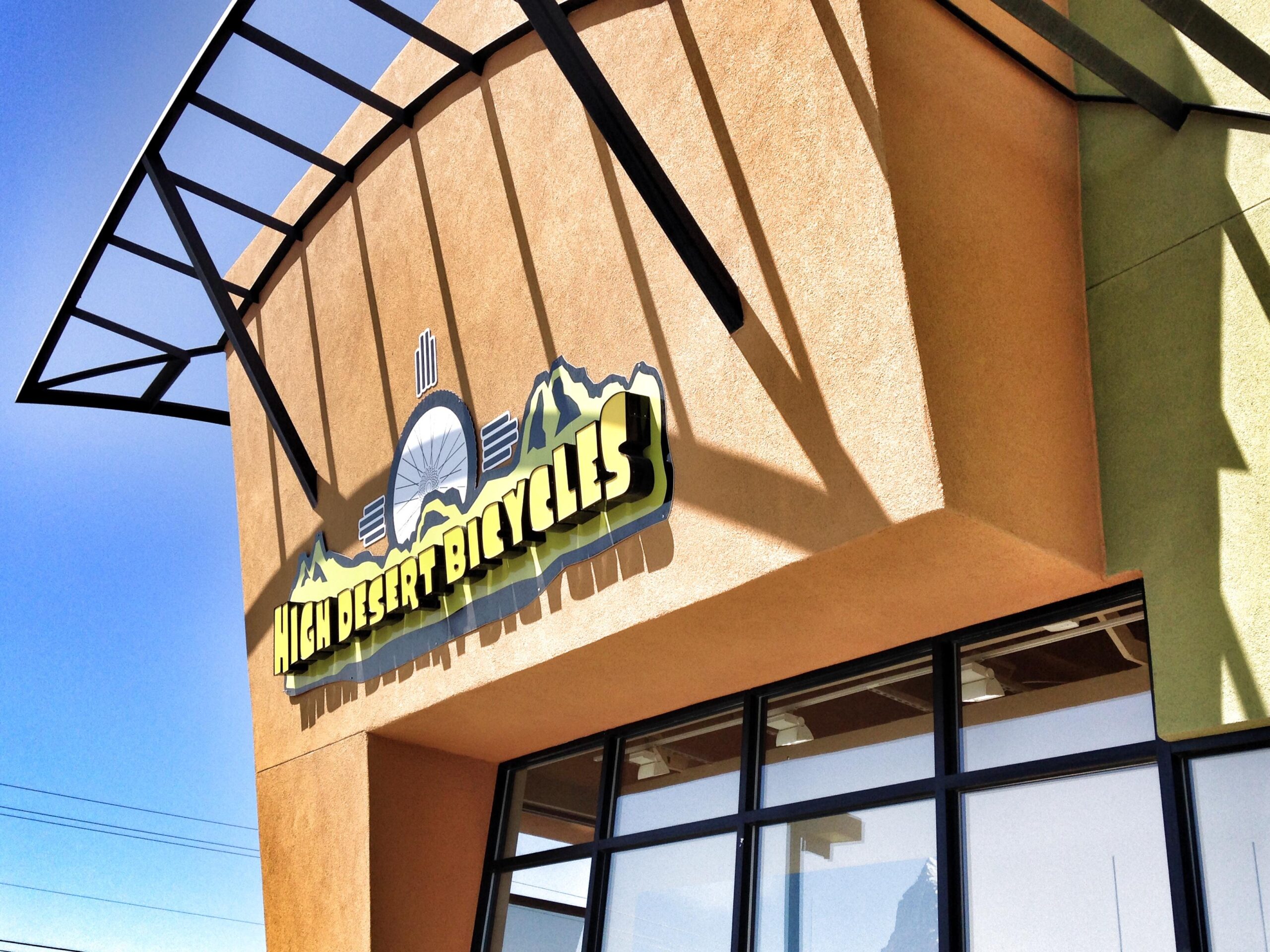Brightly lit exterior of a bicycle shop featuring a prominent sign that reads "High Desert Bicycles," with a backdrop of clear blue sky and architectural details like a black canopy casting shadows on the building's textured wall.