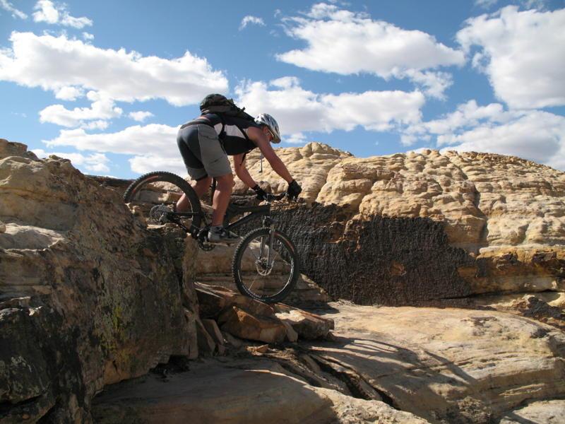 Specialized Enduro Expert: A mountain biker navigating rocky terrain under a blue sky with scattered clouds. The rider leans forward on the bike, preparing to descend a rocky ledge.