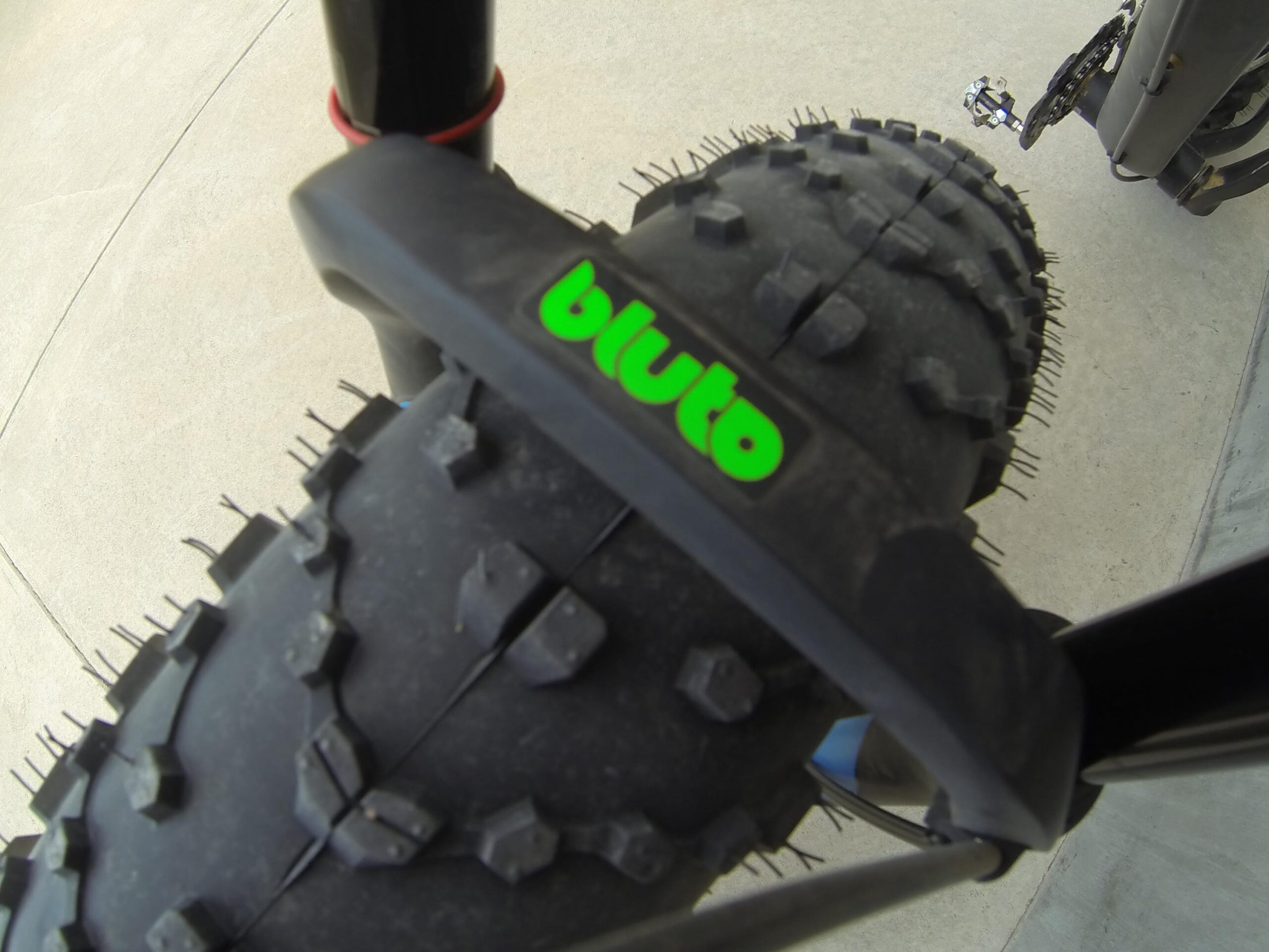 Scott Big Ed: Close-up view of a fat bike tire with a prominent "bluto" logo. The tire features a rugged tread pattern designed for traction, with visible spikes. The background shows a smooth concrete surface.