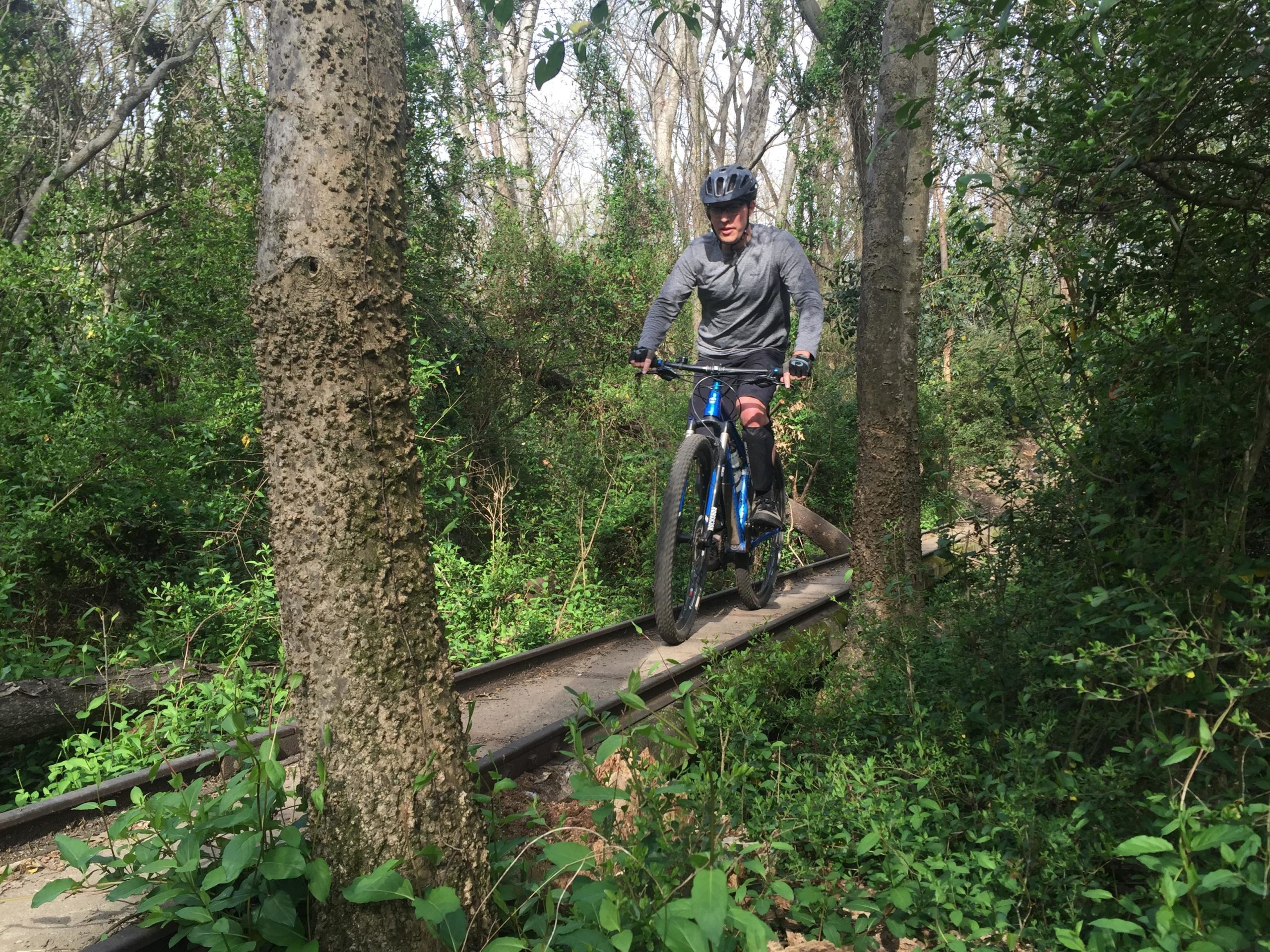 A person riding a mountain bike along a narrow, overgrown path in a wooded area, surrounded by lush greenery and trees. The rider is wearing a helmet and athletic clothing, leaning forward as they navigate over a section of track. Back Yard Trails mountain bike trail.