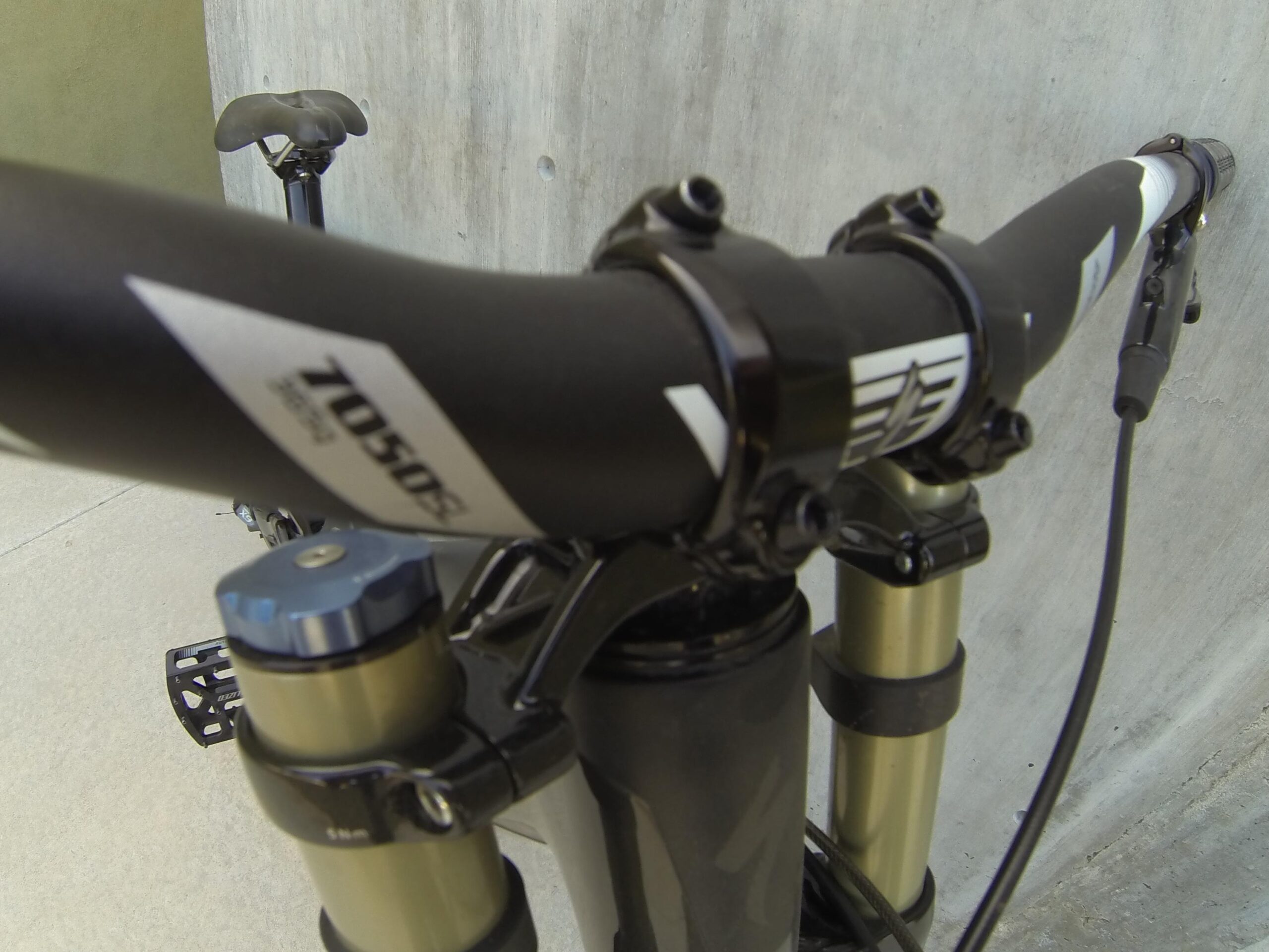 Specialized S-Works Demo 8: A close-up view of a mountain bike's handlebars and suspension system, featuring a black handlebar with silver accents and a gear shifter. The background shows a concrete surface with a bike seat slightly visible. The image highlights the bike's mechanical components and design details.