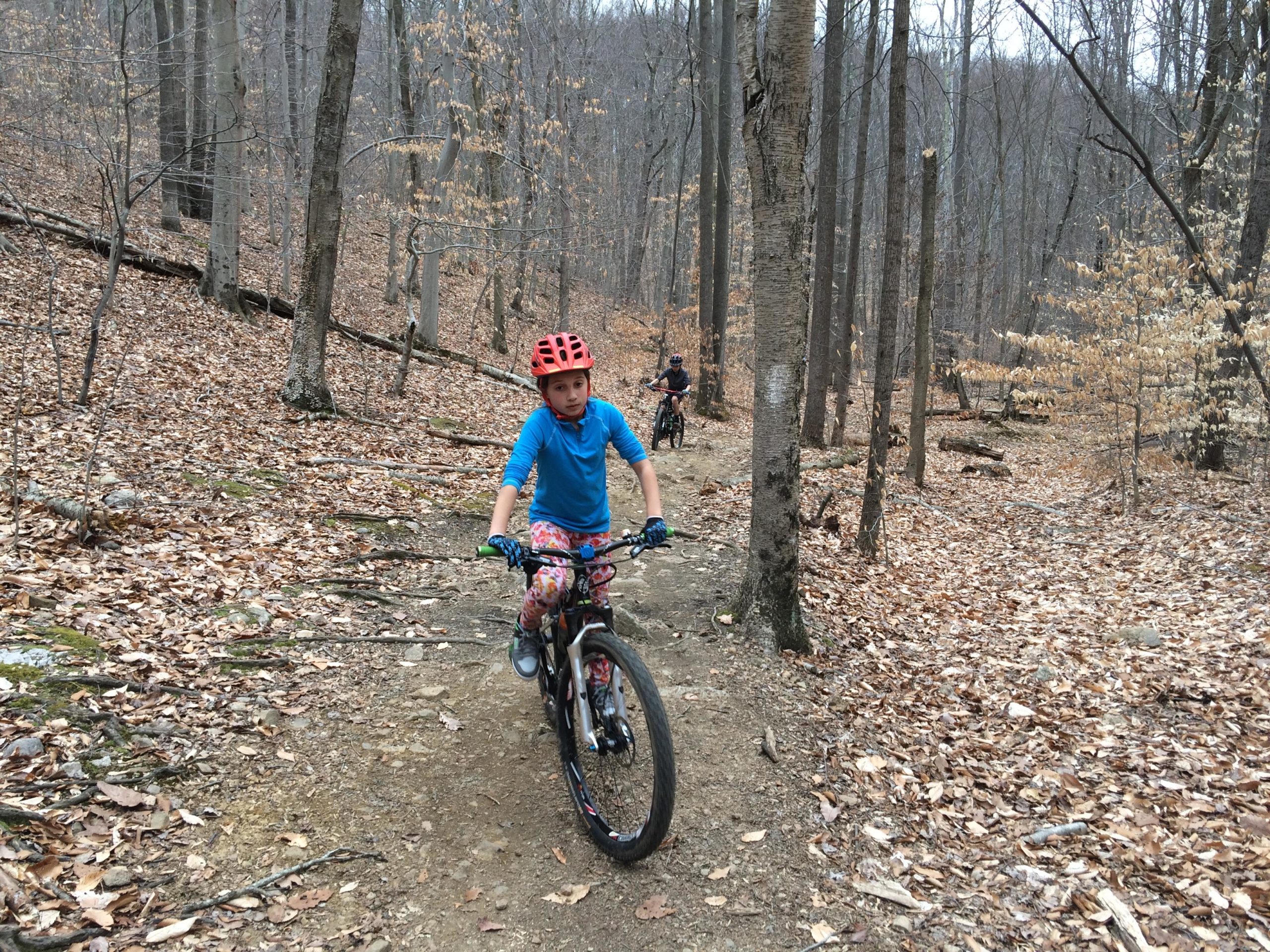 A child in a blue shirt and red helmet rides a mountain bike along a dirt trail in a wooded area, surrounded by trees and fallen leaves. Another cyclist is visible in the background. The scene is set in a natural environment during a cool season, with bare branches and dried foliage. Lewis Morris mountain bike trail.