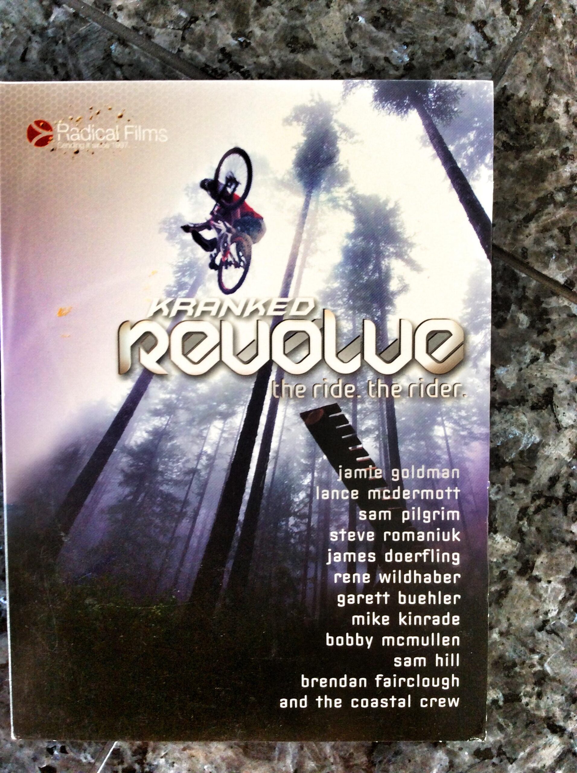 Cover of "Kranked: Revolve," featuring a mountain biker performing a trick against a backdrop of tall trees. The title and tagline "the ride. the rider." are prominently displayed, along with a list of featured riders' names. The design suggests an action-packed film focused on biking adventures.