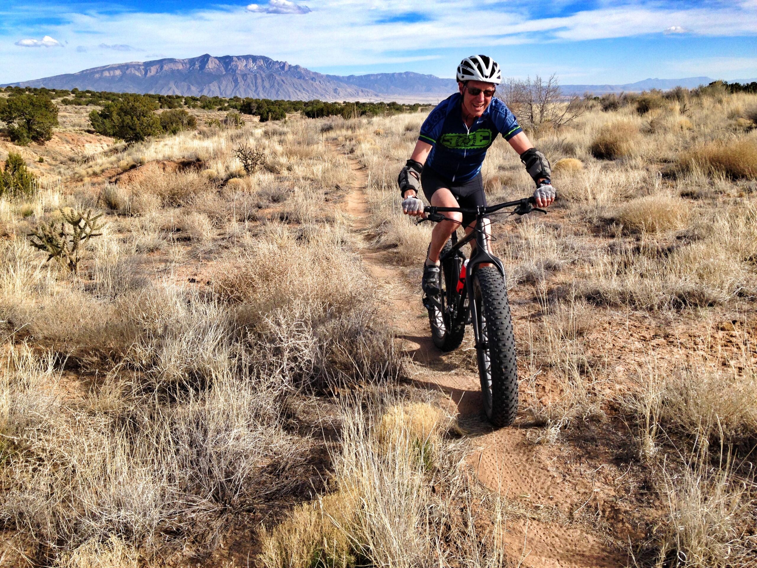 A person riding a fat bike on a dirt trail in a desert landscape, surrounded by dry grasses and shrubs, with mountains visible in the background under a partly cloudy sky. The rider is wearing a helmet and sunglasses, smiling as they navigate the trail. Parkway Fatbike trail mountain bike trail.