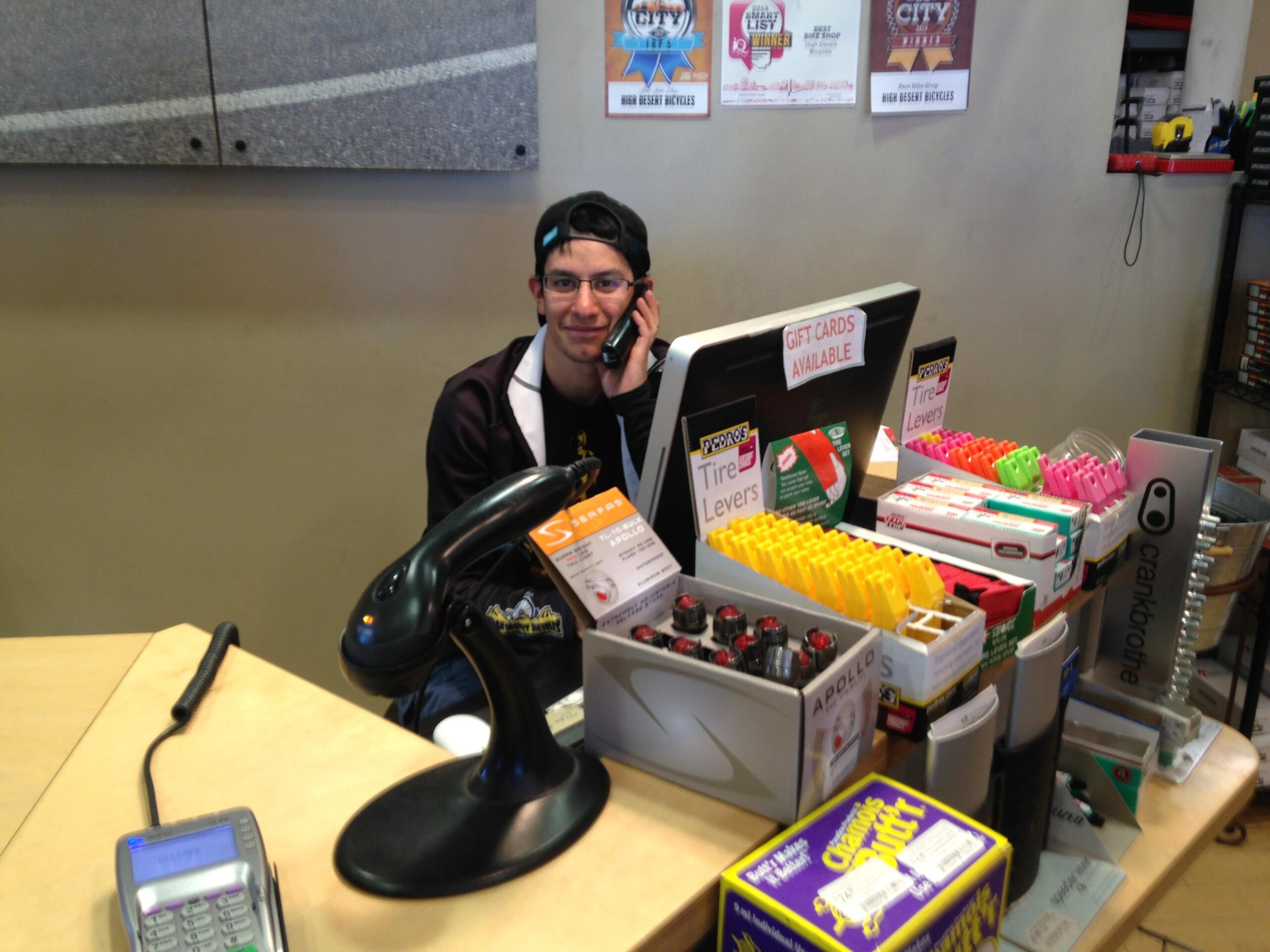 A young man wearing glasses and a baseball cap is sitting behind a retail counter, talking on a phone. The counter is stocked with various bicycle accessories, including tire levers and colorful packaging. There are also promotional signs for gift cards and bicycle products displayed on the wall behind him. A barcode scanner and a cash register are visible on the counter.
