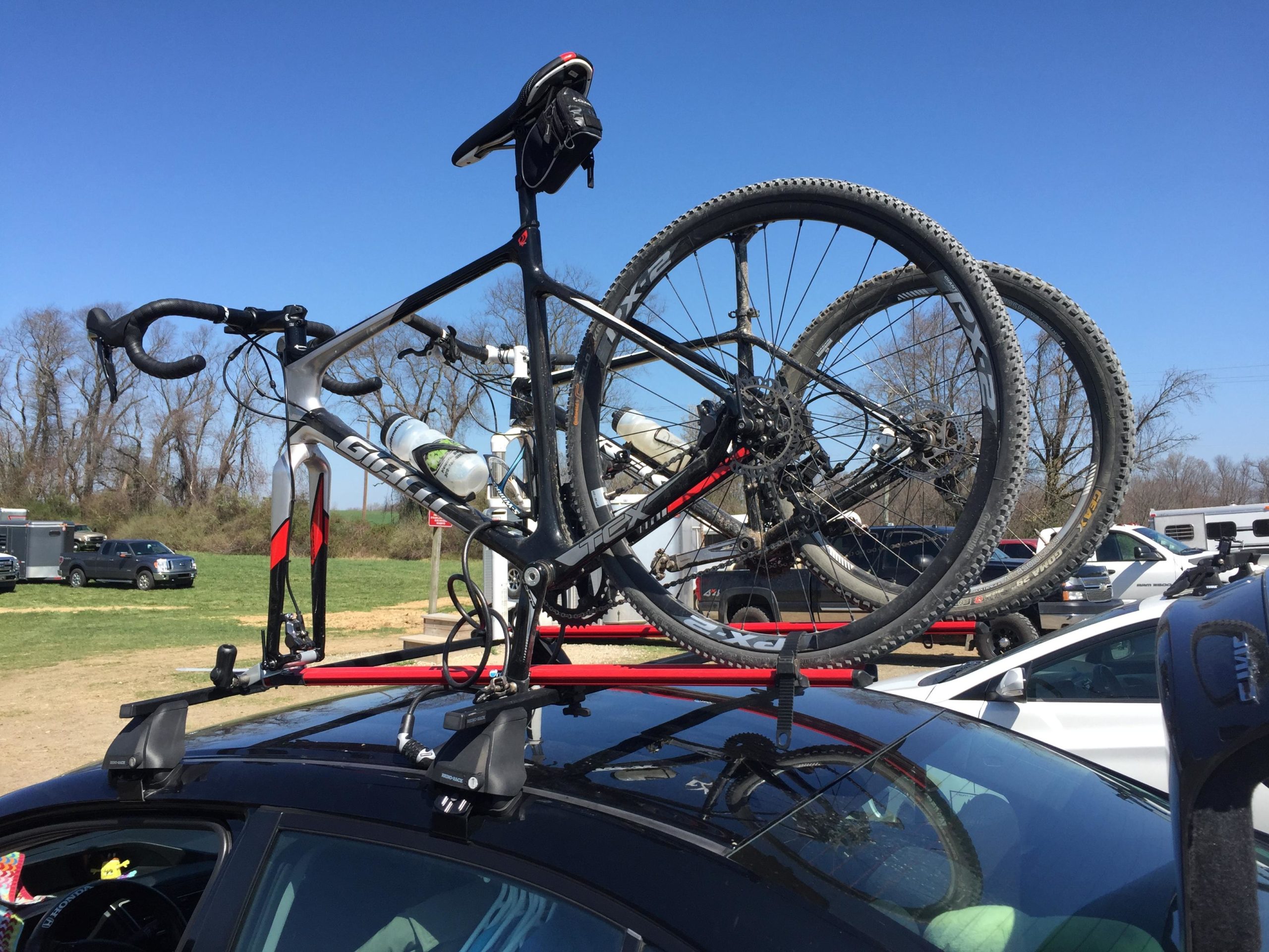 A bicycle is mounted on a roof rack of a car, with two tires visible. The background features a clear blue sky and a parking area with vehicles. The scene suggests outdoor recreational activities. Fair Hill mountain bike trail.