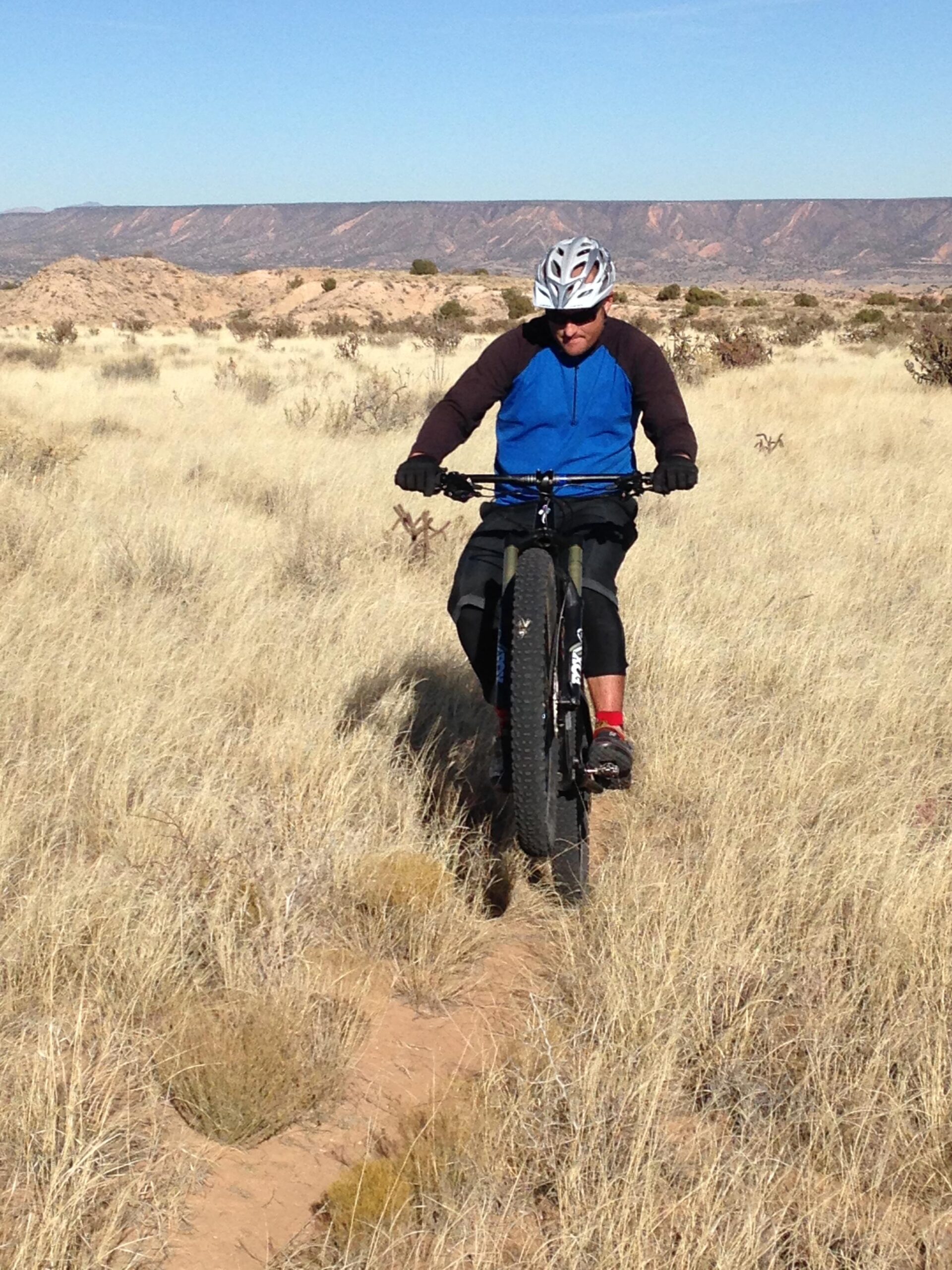 Specialized S-Works Stumpjumper FSR Carbon: A cyclist in a blue long-sleeve shirt and black shorts rides a fat bike on a dirt trail through tall, dry grass with a mountainous landscape in the background under a clear blue sky.