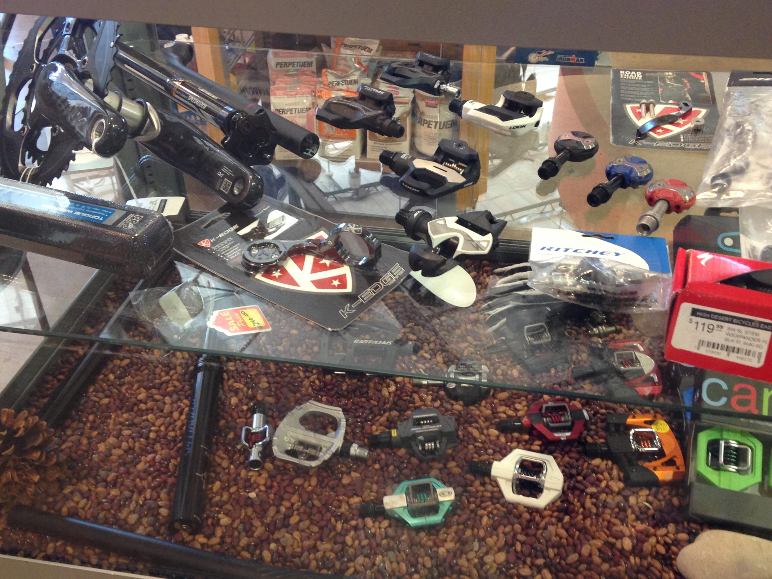 A display case filled with various bicycle components and accessories, including pedals, cranksets, and tools, arranged over a bed of coffee beans. Some items are packaged and labeled with prices, showcasing brands like Ritchey and K-Edge.