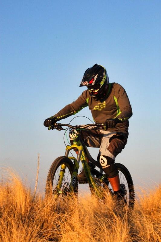 A mountain biker wearing a helmet and protective gear navigates a grassy hillside at sunset, focusing intently on the trail ahead. The sky is clear and blue, creating a vibrant backdrop for the action.