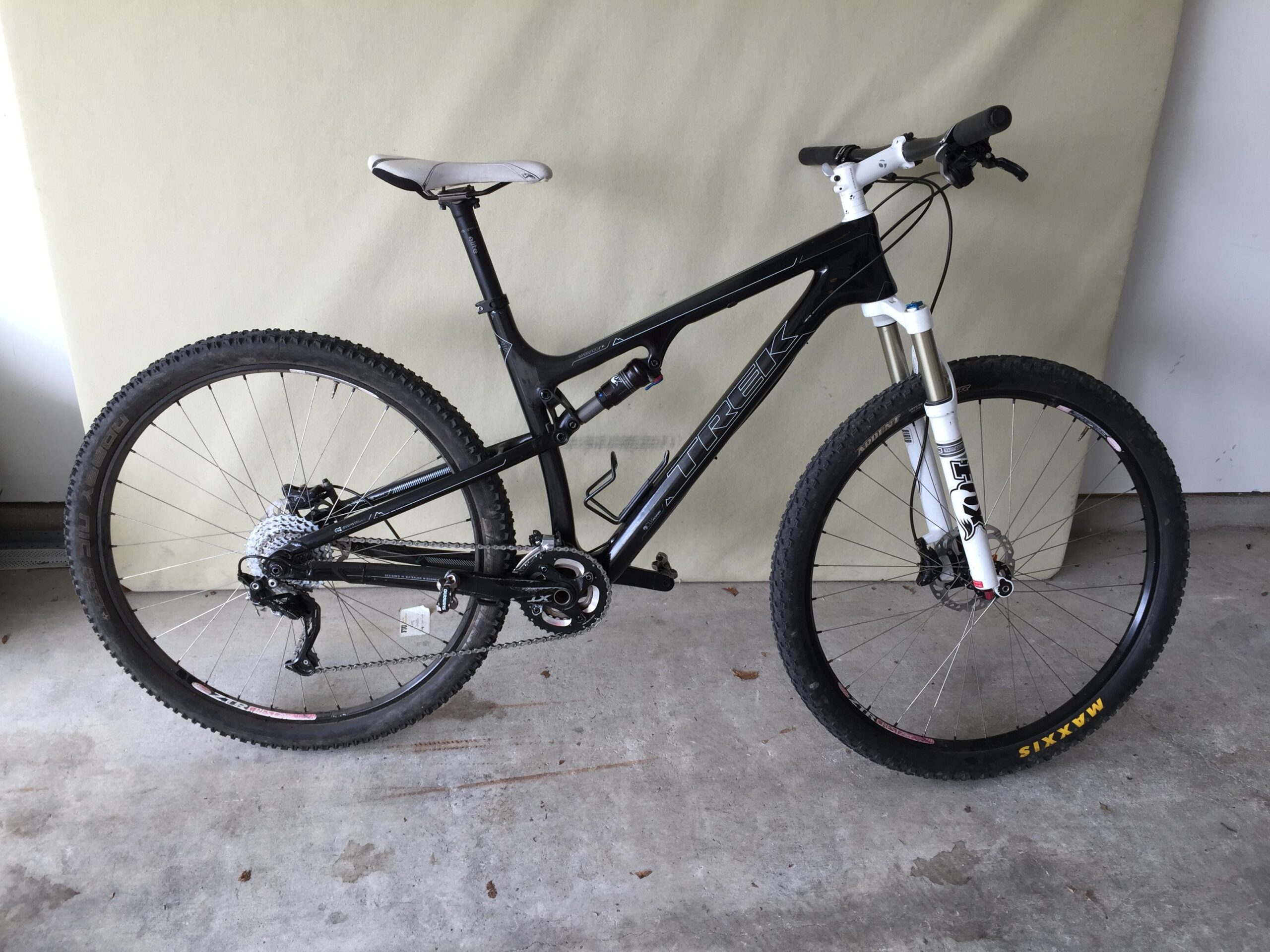 Trek Superfly 100 AL: A black and white mountain bike is positioned upright on a concrete surface against a light green backdrop. The bike features thick tires, a sturdy frame, and a suspension fork, indicating it is designed for off-road riding. The drivetrain and gear set are visible, showcasing a high-performance setup.