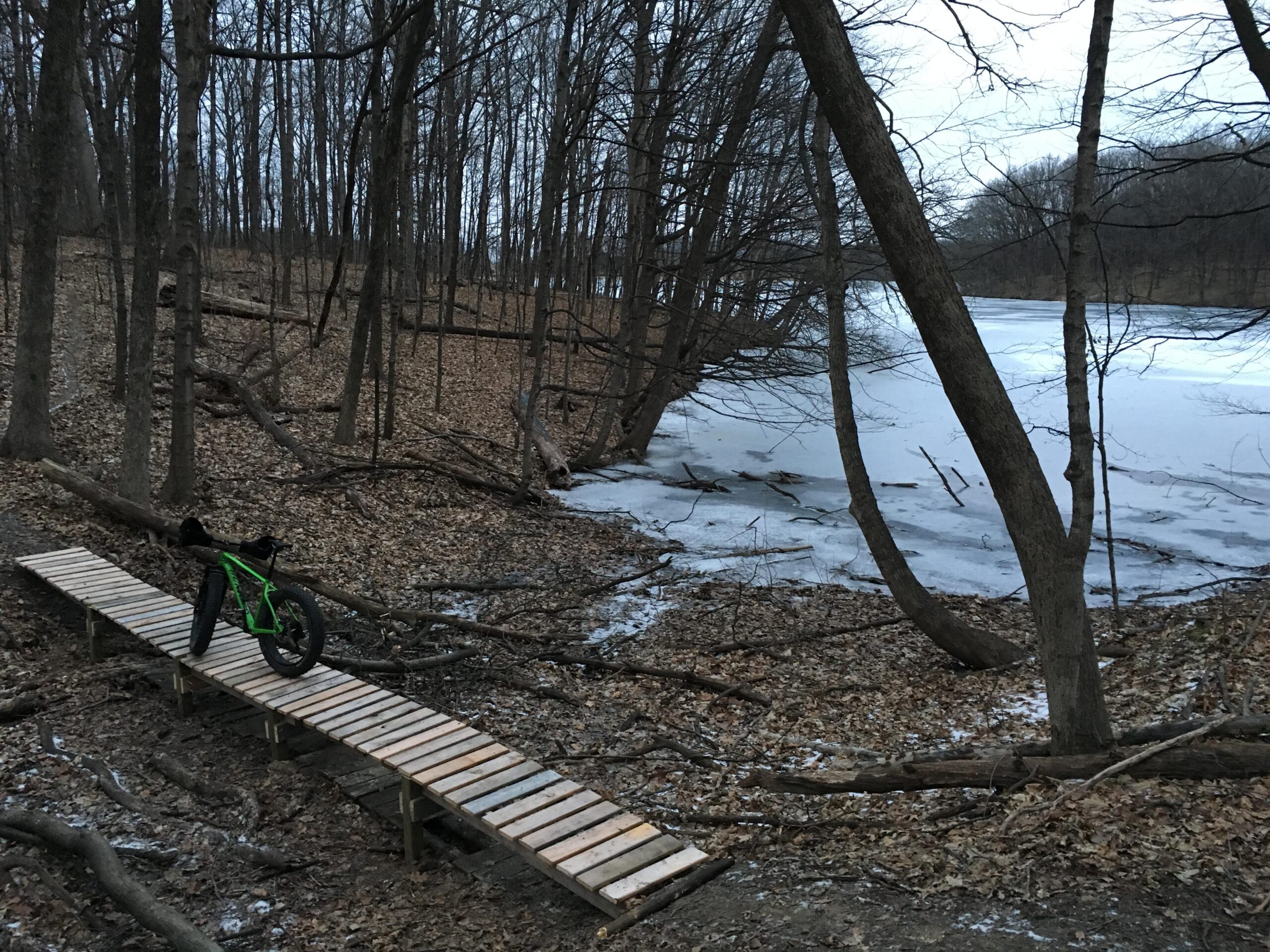 A green fat bike is positioned on a wooden plank bridge that extends over a frozen body of water, surrounded by leafless trees and a forest floor covered with dry leaves and branches. The scene is set during twilight, creating a calm and serene atmosphere in the woods. Lake Storey Trail System mountain bike trail.