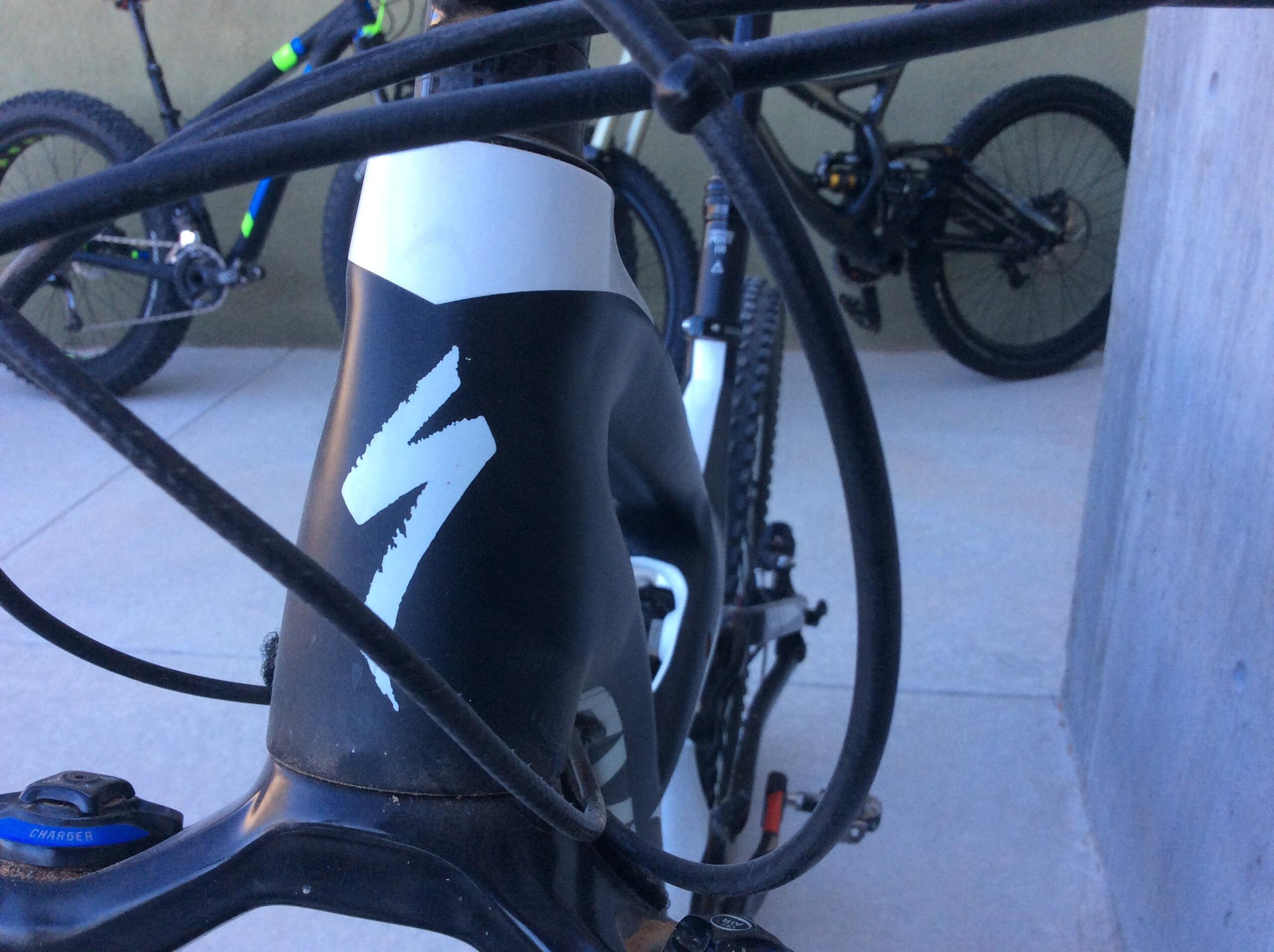 Close-up view of a mountain bike frame with a partially visible logo, surrounded by bike cables. In the background, another mountain bike is faintly visible. The setting appears to be an outdoor area with a smooth concrete surface.