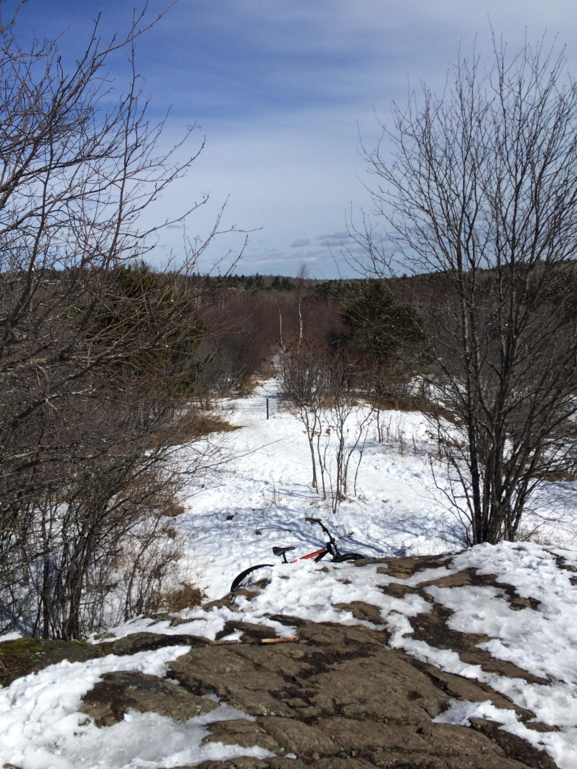 A scenic winter landscape featuring a rocky outcrop with patches of snow. In the foreground, a mountain bike rests on the snow-covered ground. The background shows a winding trail leading through a forest with bare trees and a clear blue sky above. Hartley Park mountain bike trail.