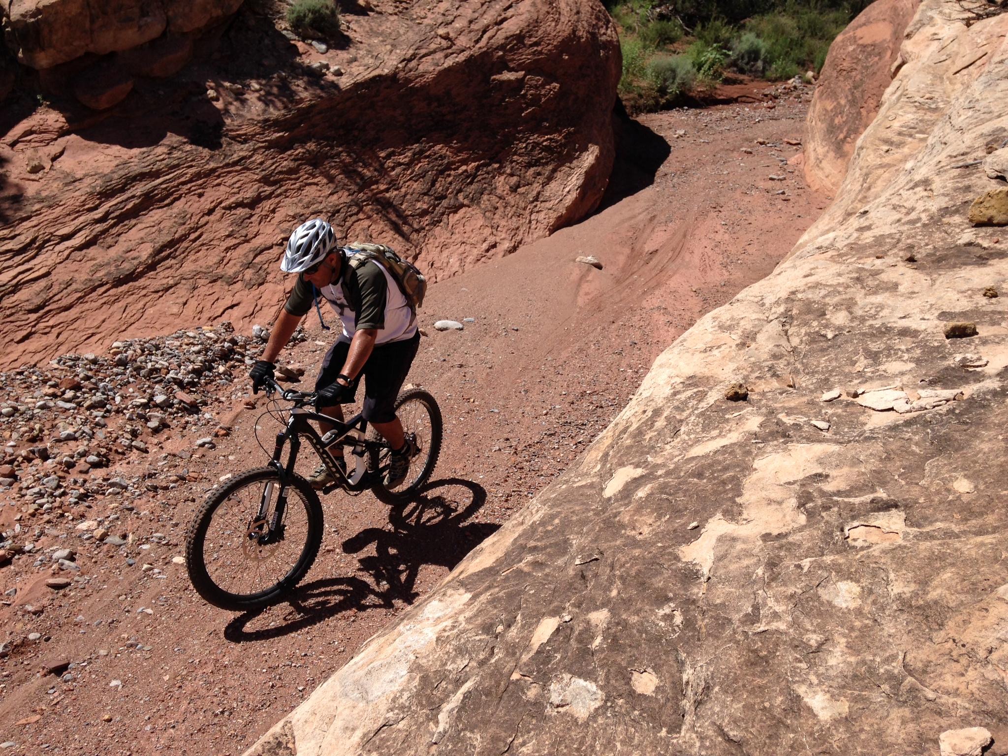 Specialized Enduro S-Works 650b: A person riding a mountain bike on a rocky, desert trail surrounded by reddish-brown rock formations and sparse vegetation. The cyclist is wearing a helmet, gloves, and a backpack, focusing on navigating the uneven terrain.