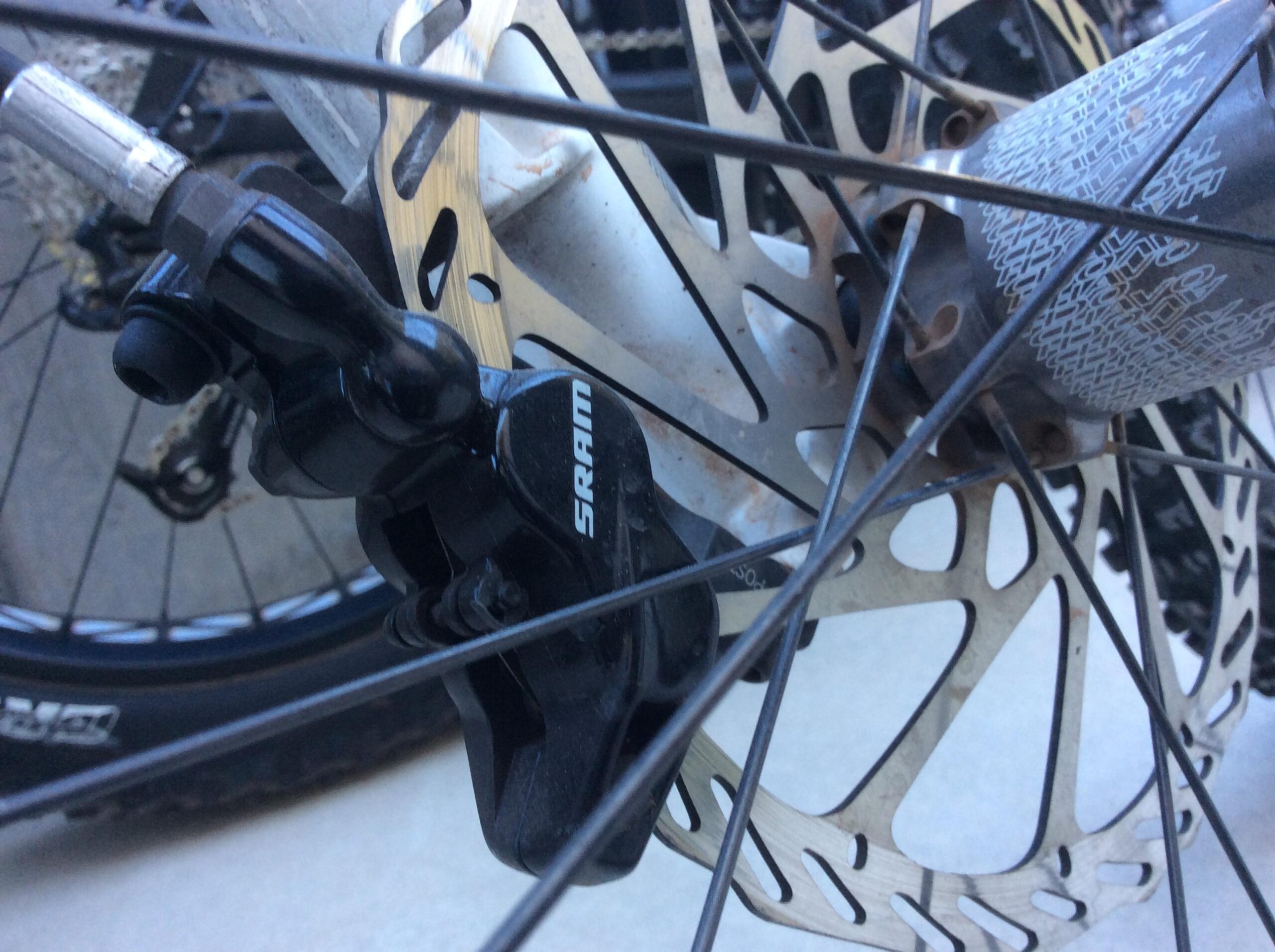 Close-up of a SRAM disc brake caliper mounted on a bicycle wheel, featuring a metal rotor with a distinctive pattern and wheel spokes in the background.