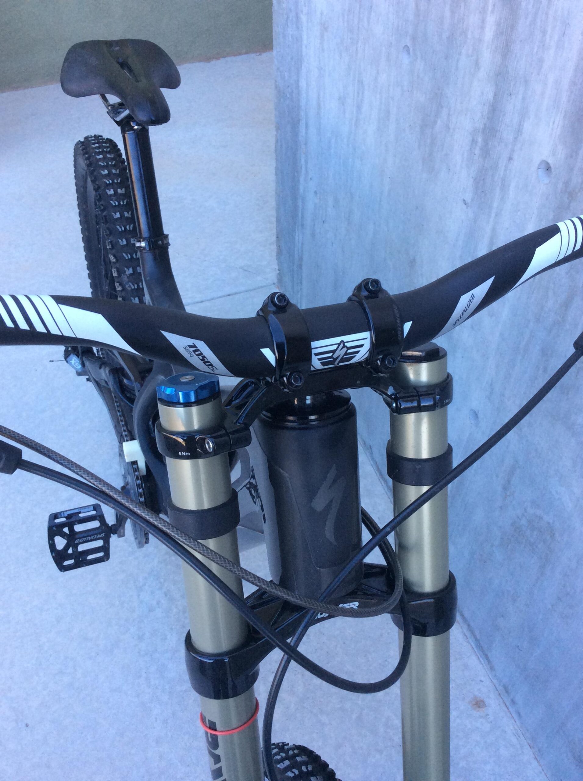 Specialized S-Works Demo 8 Carbon: Close-up view of a mountain bike's handlebars and front suspension, featuring a black and white handlebar, adjustable suspension components, and visible brake cables. The background shows a smooth, light-colored surface and a concrete wall.