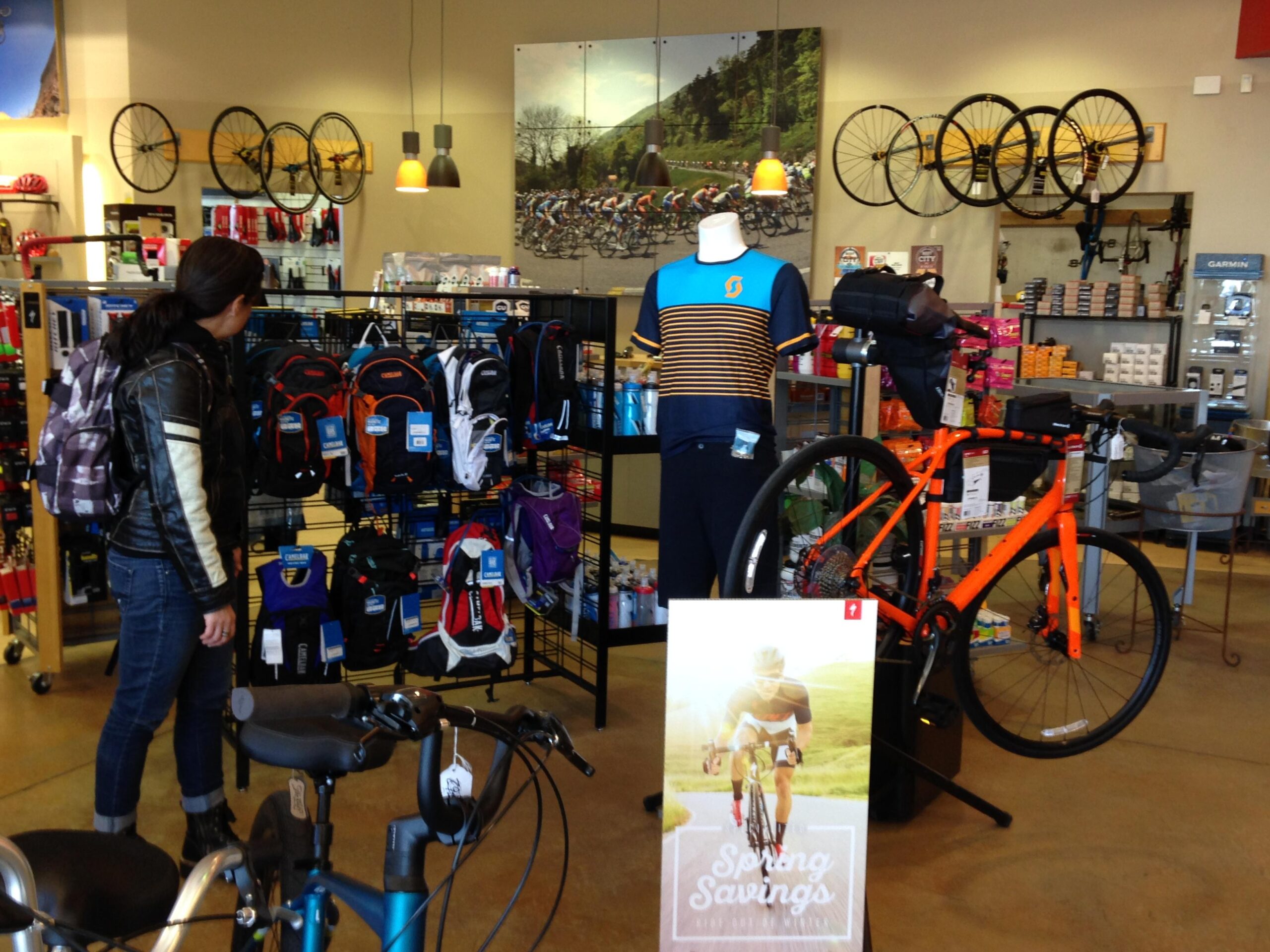 A retail bike shop interior featuring a woman examining a variety of backpacks hanging on a rack, while an orange bicycle is prominently displayed beside her. The background includes cycling-related merchandise, wall decor with cycling images, and a promotional sign advertising "Spring Savings."