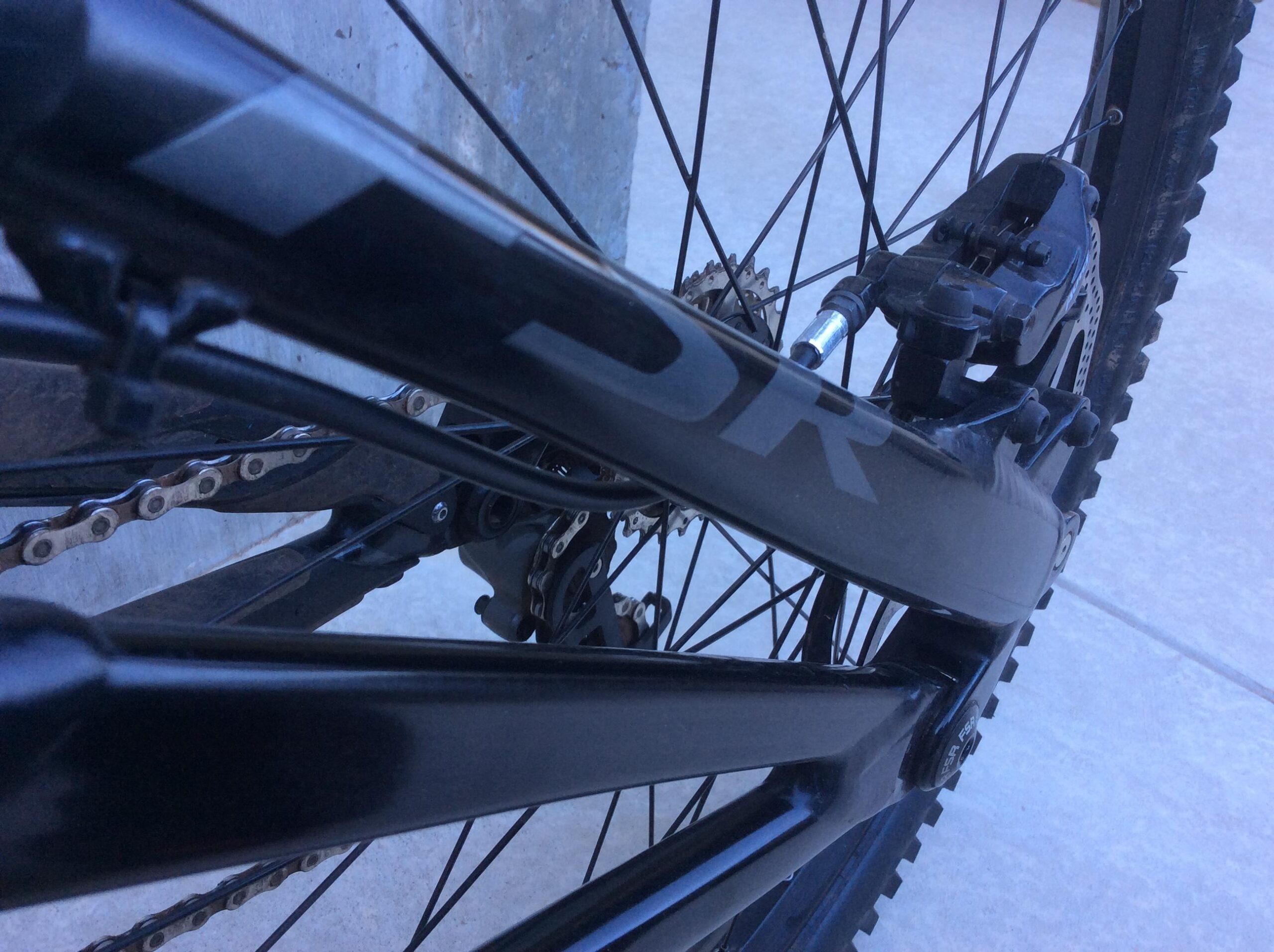 Specialized S-Works Demo 8 Carbon: Close-up view of a black mountain bike frame, highlighting the chain, derailleur, and wheel components. The image emphasizes the bike's structural design and mechanics against a light concrete background.