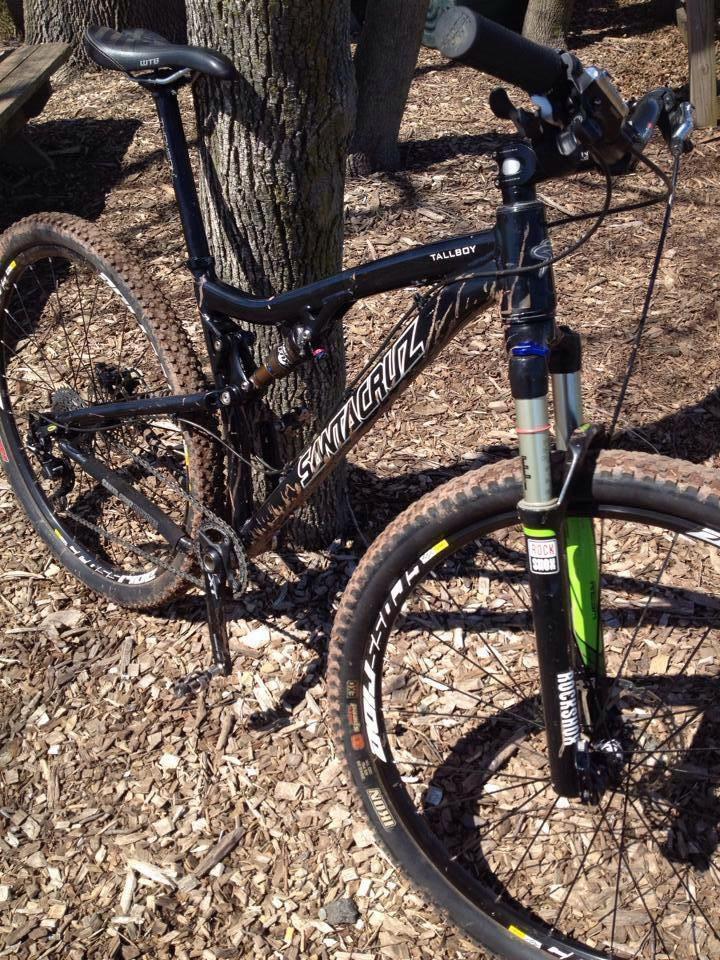 A black Santa Cruz Tallboy mountain bike leaning against a tree, with a focus on its front suspension fork and knobby tires, positioned on a bed of wood chips. Lewis Morris mountain bike trail.